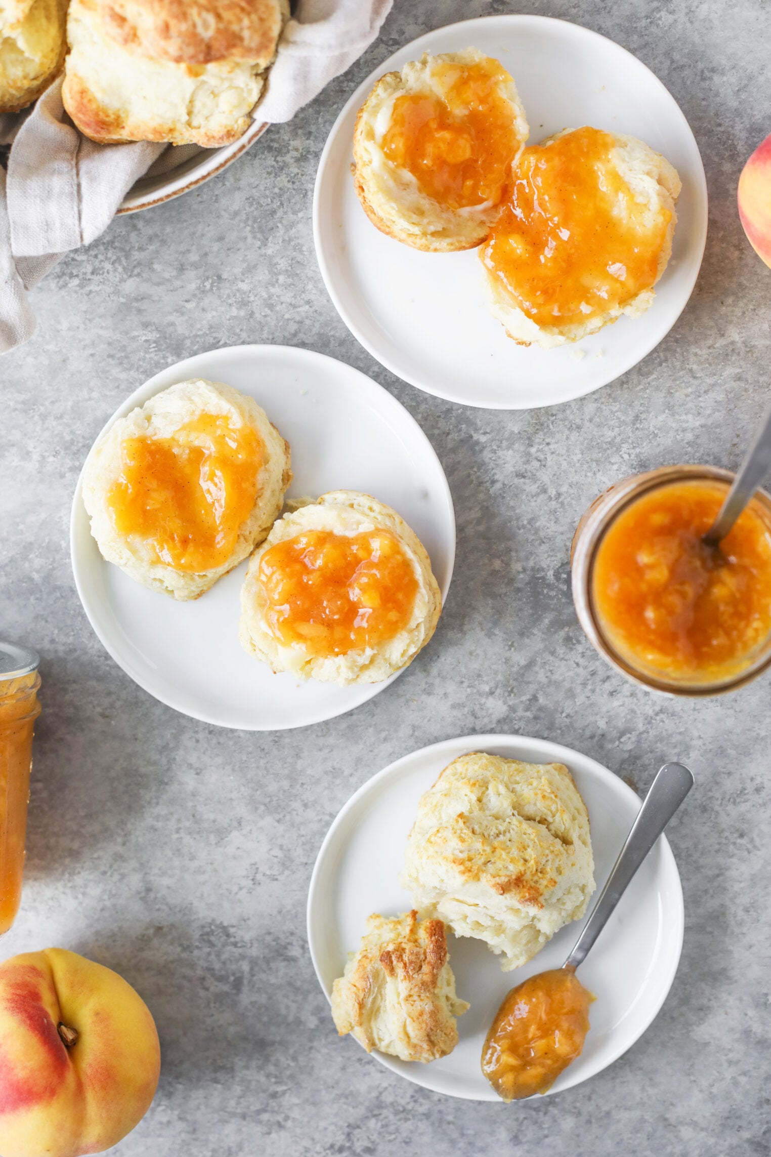 Three White Plates With Biscuits Topped With Low Sugar Peach Jam, A Jar Of Jam With A Spoon, Fresh Peaches, And More Biscuits In A Basket Are Arranged On A Light Gray Surface.