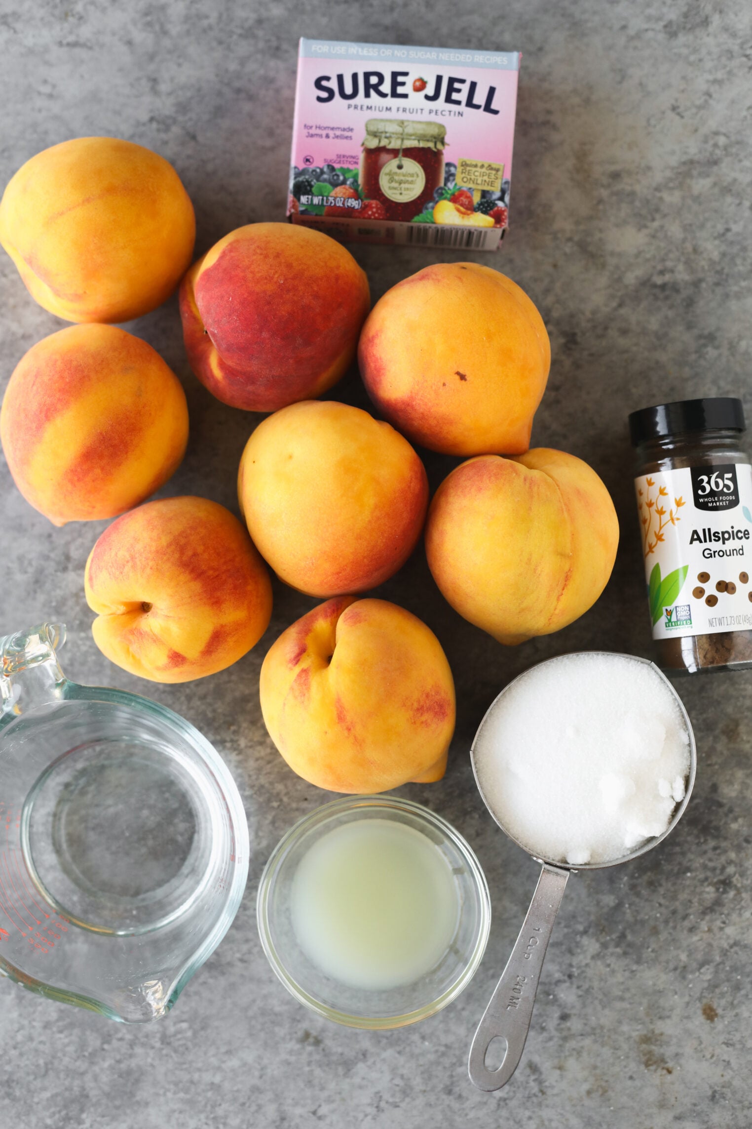 Peaches, A Box Of Sure-Jell Pectin, A Jar Of Ground Allspice, A Glass Measuring Cup With Water, Lemon Juice In A Small Bowl, And A Cup Of Sugar Are Arranged On A Gray Countertop—Everything You Need To Make Delicious Low Sugar Peach Jam.