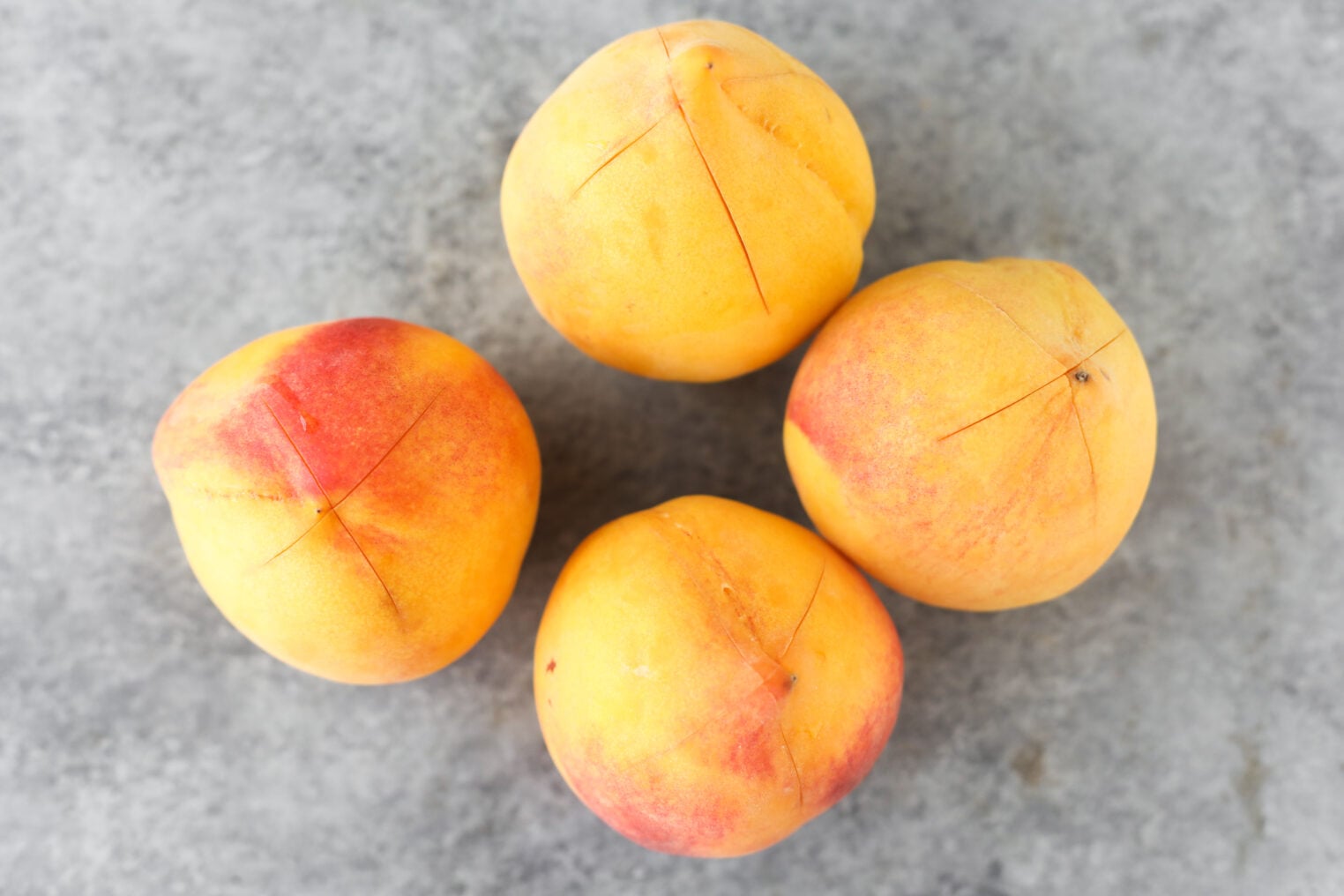 Four Ripe Peaches With Reddish And Yellow Skin Sit On A Gray Textured Surface. Each Peach Has A Shallow X-Shaped Cut On Top, Likely To Help With Peeling—Perfect Preparation For Making Low Sugar Peach Jam.
