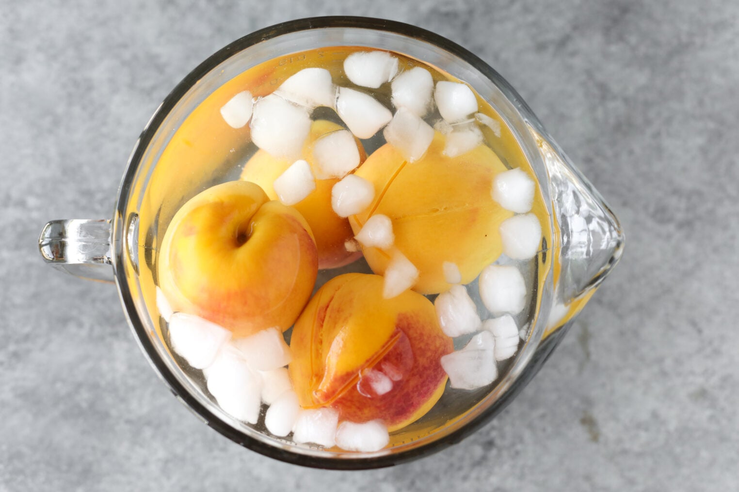Three Peaches, Perfect For Making Low Sugar Peach Jam, Are Submerged In A Glass Pitcher Filled With Ice Water And Ice Cubes, Set On A Gray Surface.