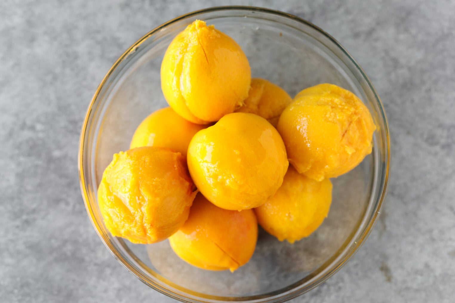 A Glass Bowl Filled With Several Peeled, Whole Yellow Peaches—Perfect For Making Low Sugar Peach Jam—Is Placed On A Gray Marbled Surface.