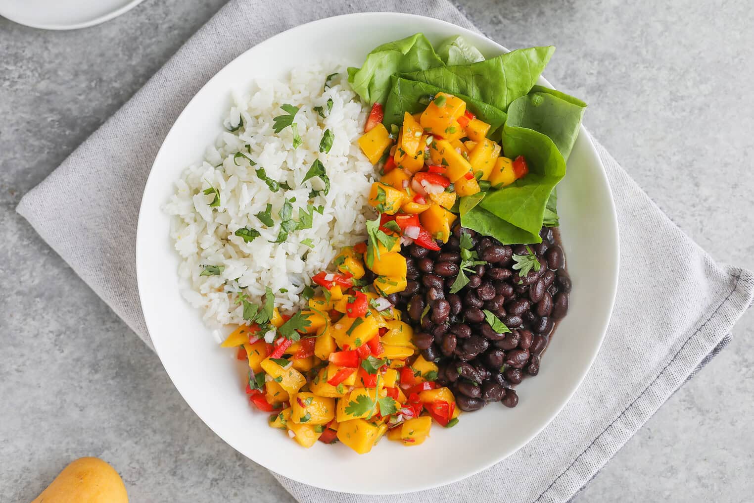 A White Bowl Featuring White Rice With Herbs, Black Beans, Vibrant Mango Salsa With Red Peppers, And Fresh Spinach Leaves—Perfect For Delicious Mango Salsa Bowls—Rests On A Gray Cloth On A Gray Surface.