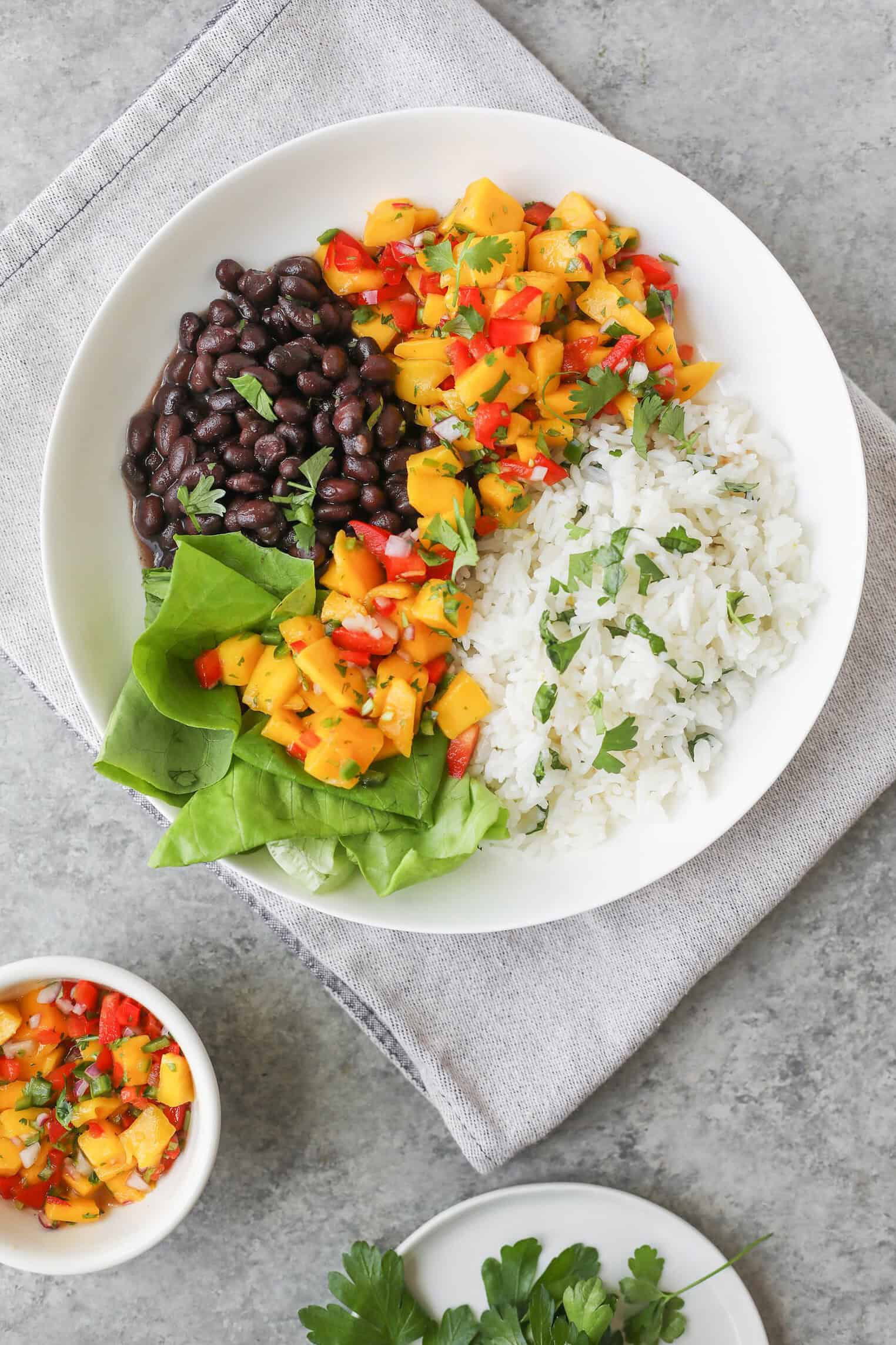 A White Bowl With Black Beans, White Rice With Herbs, Leafy Greens, And Mango Salsa Creates Vibrant Mango Salsa Bowls, Set On A Gray Napkin. Small Bowls Of Salsa And Fresh Herbs Sit Nearby On A Light Gray Surface.