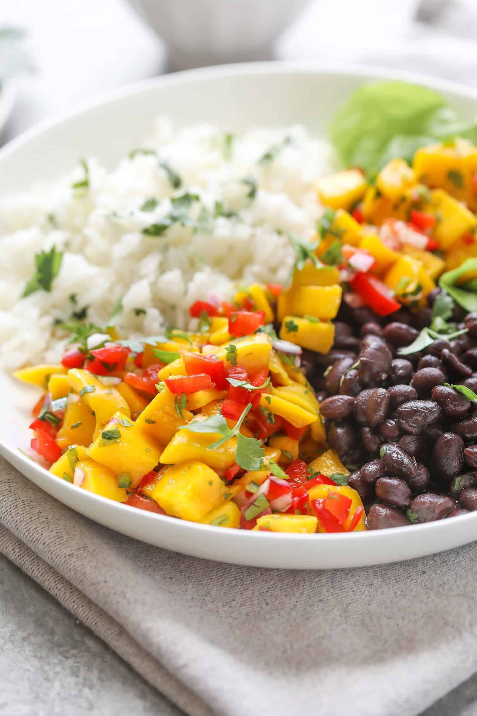 A White Bowl Filled With White Rice, Black Beans, And Vibrant Mango Salsa Bowls Made With Diced Mango, Red Bell Pepper, And Fresh Herbs, All Served On A Neutral Napkin.