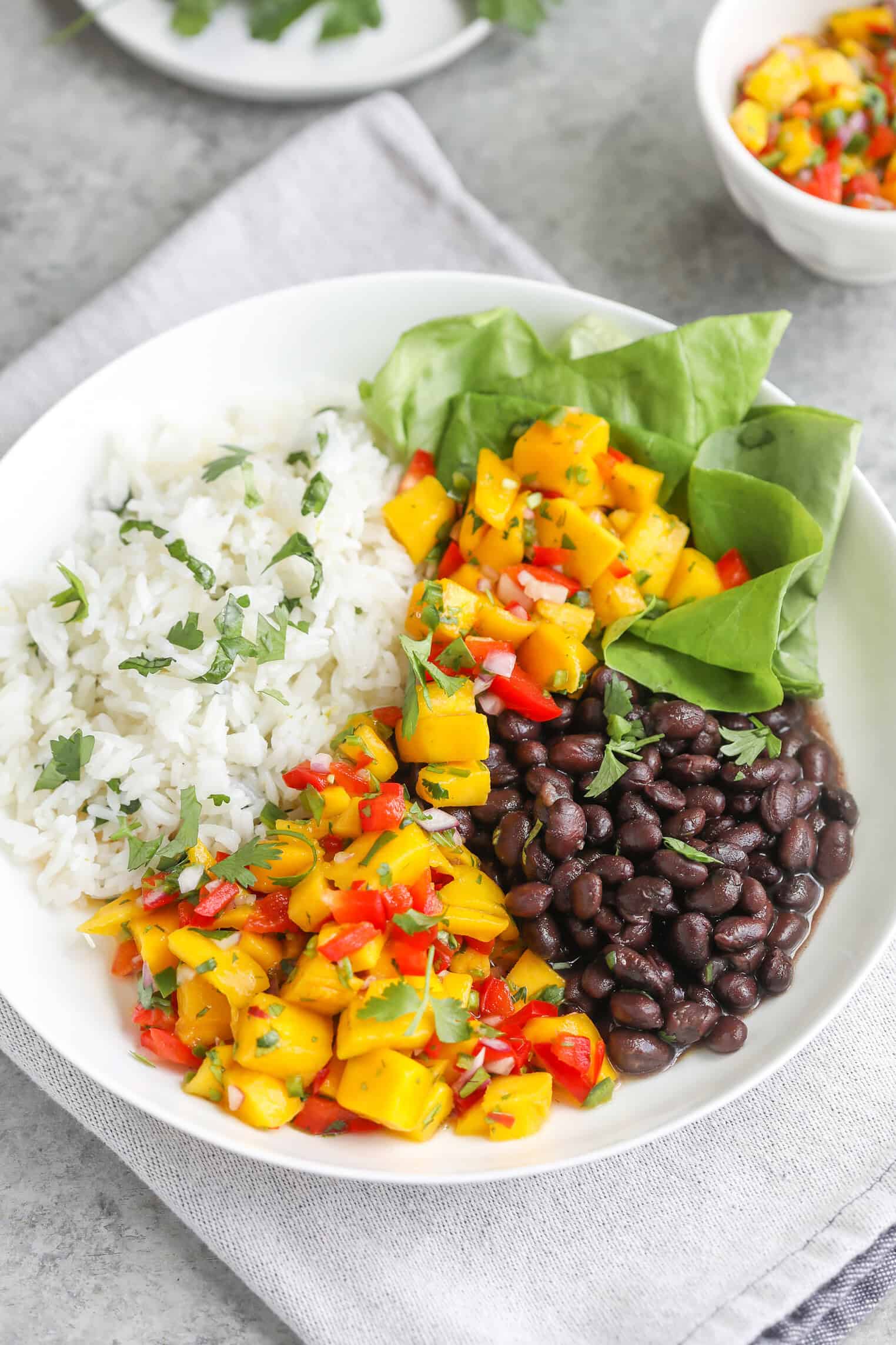 A White Bowl Features A Vibrant Mango Salsa Rice Bowl—White Rice, Black Beans, Spinach Leaves, And Zesty Mango Salsa With Diced Mango, Red Pepper, Onion, And Herbs—All Served On A Light Gray Napkin.