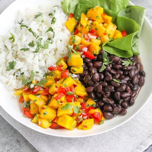 A White Bowl Features A Vibrant Mango Salsa Rice Bowl—White Rice, Black Beans, Spinach Leaves, And Zesty Mango Salsa With Diced Mango, Red Pepper, Onion, And Herbs—All Served On A Light Gray Napkin.