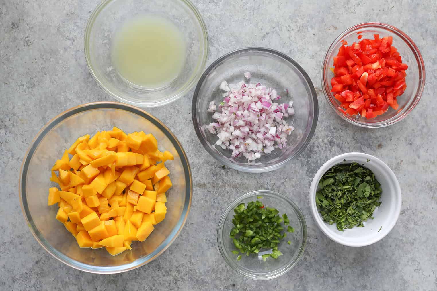 Six Small Bowls On A Gray Countertop Show The Fresh Ingredients For A Mango Salsa Recipe: Chopped Mango, Diced Red Onion, Bell Pepper, Cilantro, Jalapeño, And Lime Juice.