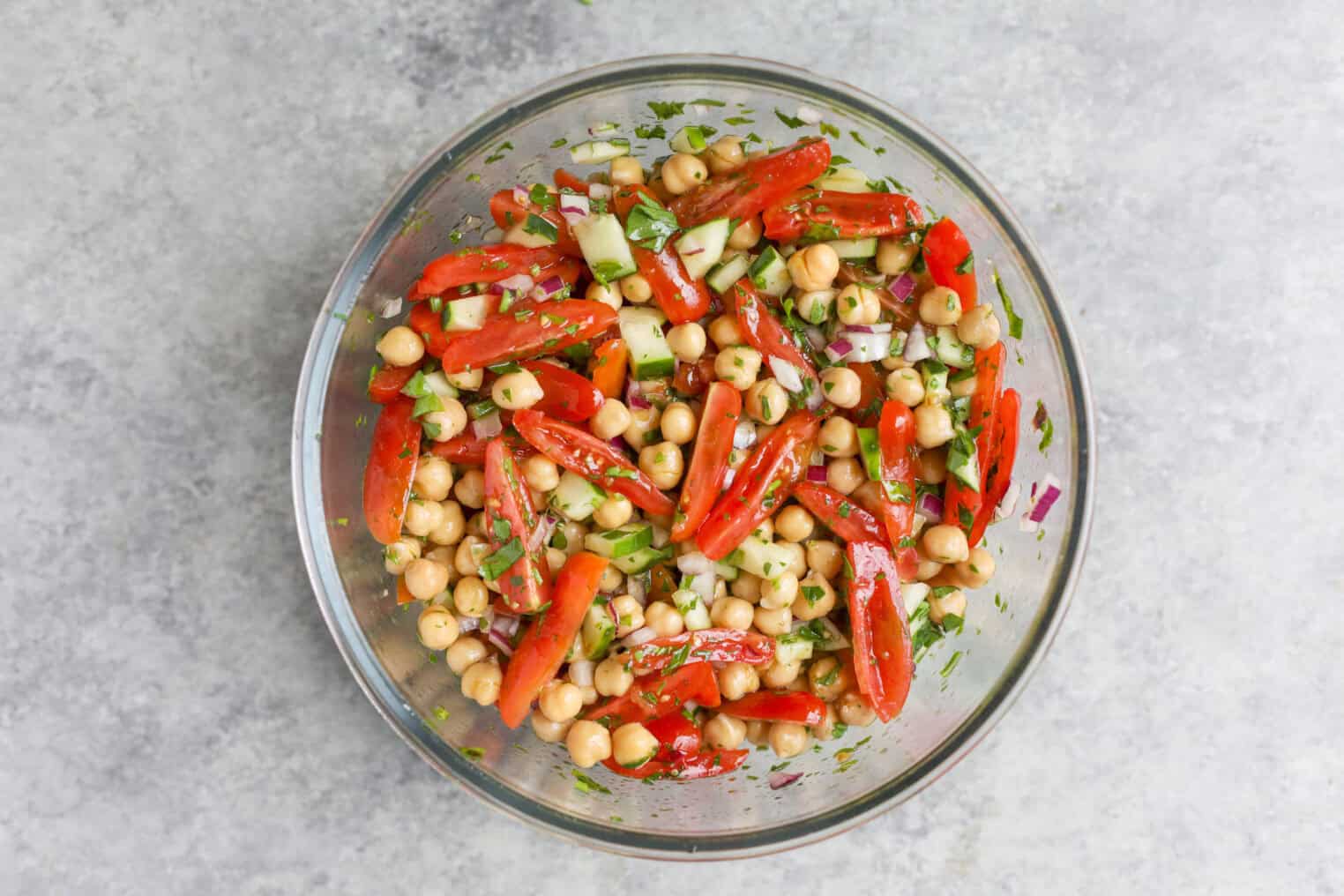 Marinated Chickpea Salad 5 A Glass Bowl Filled With Vibrant Chickpea Salad, Featuring Chopped Cucumber, Tomato Slices, Red Onion, Parsley, And Fresh Herbs, All Mixed Together On A Light Gray Surface.