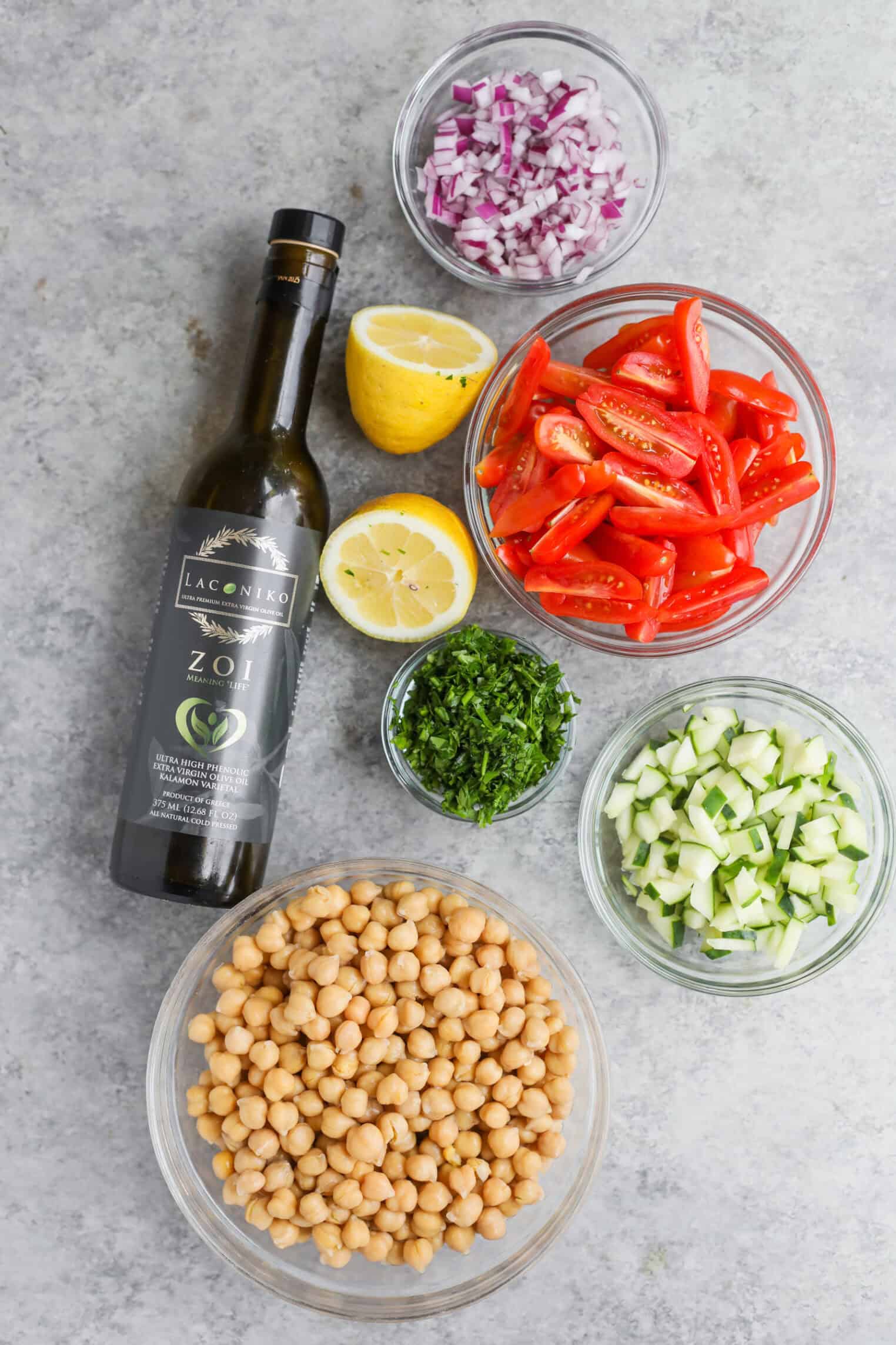 Marinated Chickpea Salad 2 A Bottle Of Olive Oil, Halved Lemon, Bowls Of Chopped Red Onion, Parsley, Cucumber, Grape Tomatoes, And A Bowl Of Chickpeas Are Arranged On A Gray Surface—Perfect For Preparing A Fresh Chickpea Salad.