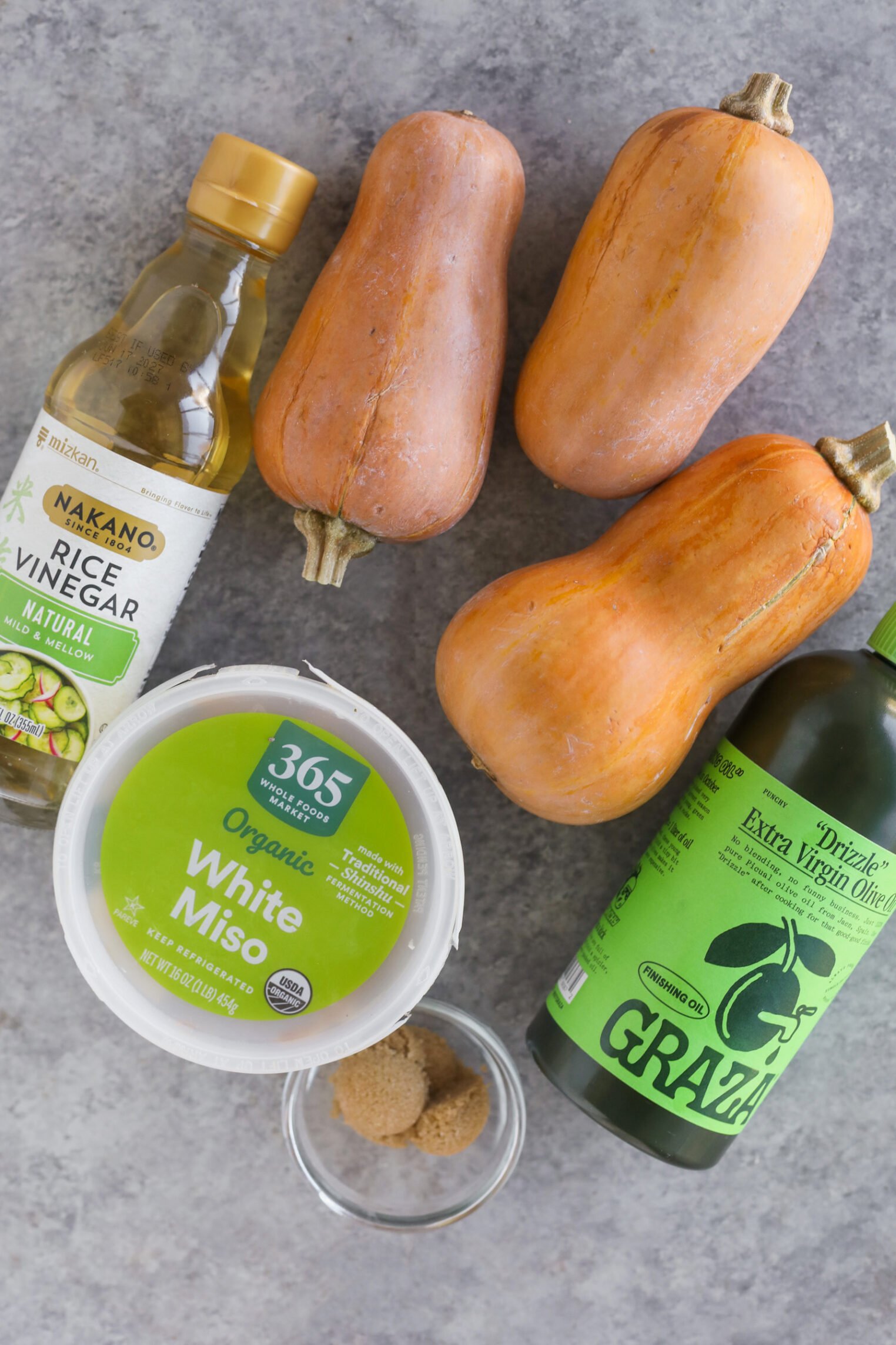 Miso Roasted Honeynut Squash 2 Three Small Butternut Squashes, Perfect For Roasted Honeynut Squash, A Bottle Of Rice Vinegar, Organic White Miso, Green Olive Oil, And A Dish Of Brown Sugar Are Arranged On A Gray Surface.