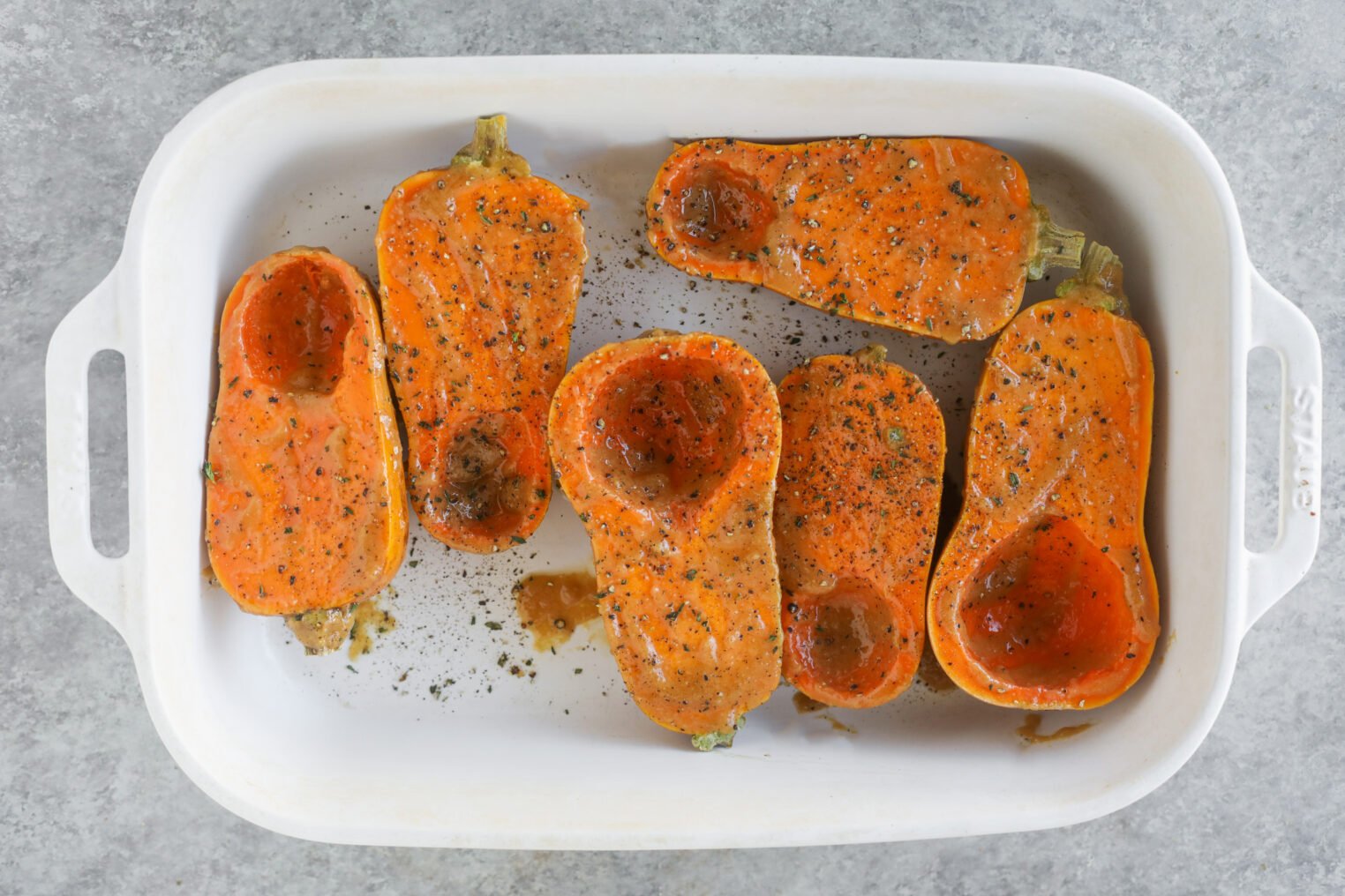 Miso Roasted Honeynut Squash 6 Roasted Honeynut Squash Halves, Seasoned With Pepper And Spices, Are Arranged Cut-Side Up In A White Ceramic Baking Dish On A Gray Surface.