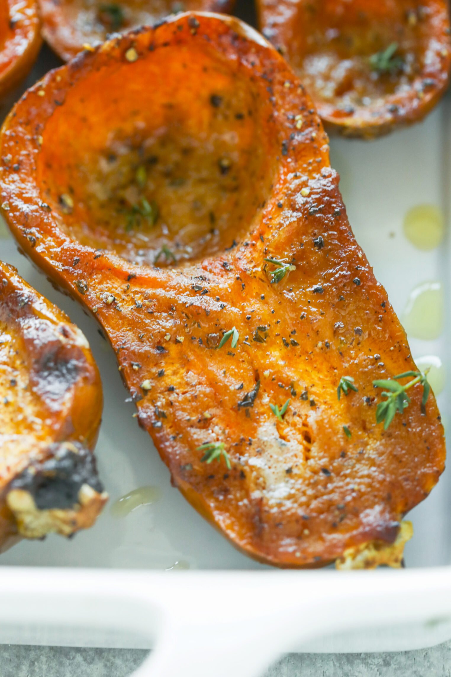 Miso Roasted Honeynut Squash 8 A Close-Up Of A Roasted Honeynut Squash Half In A White Dish, Seasoned With Herbs And Pepper, And Drizzled Lightly With Oil. Fresh Thyme Leaves Garnish The Surface.