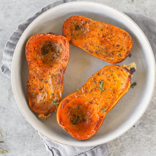 Miso Roasted Honeynut Squash 10 Three Halves Of Roasted Honeynut Squash With Herbs And Seasoning Served On A White Plate, Placed On A Gray Napkin Atop A Light Gray Surface.