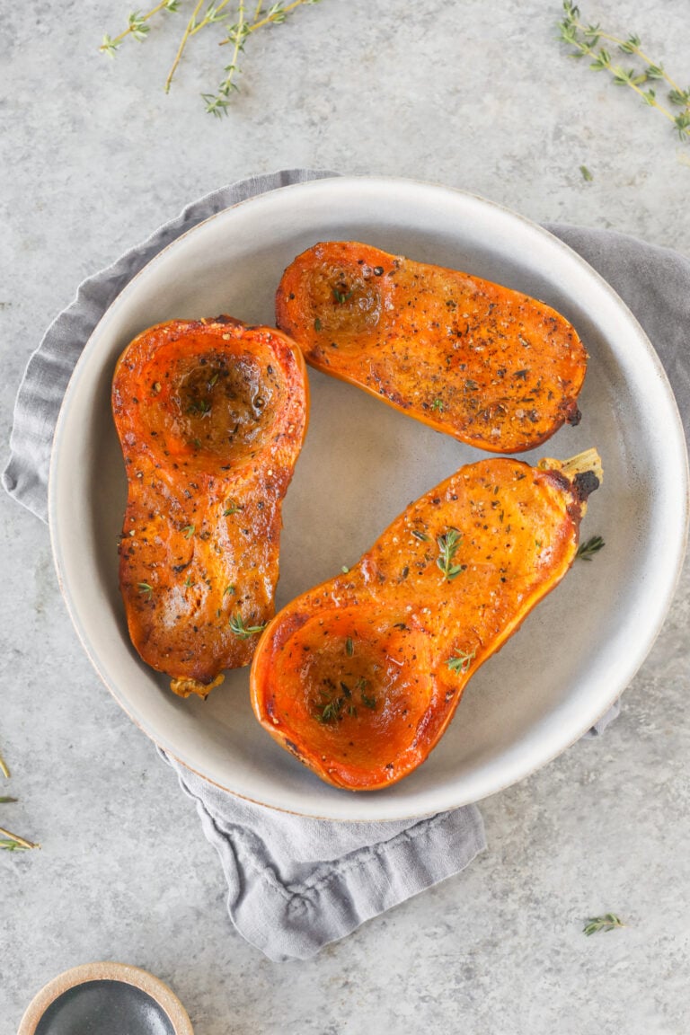 Three Halves Of Roasted Honeynut Squash With Herbs And Seasoning Served On A White Plate, Placed On A Gray Napkin Atop A Light Gray Surface.