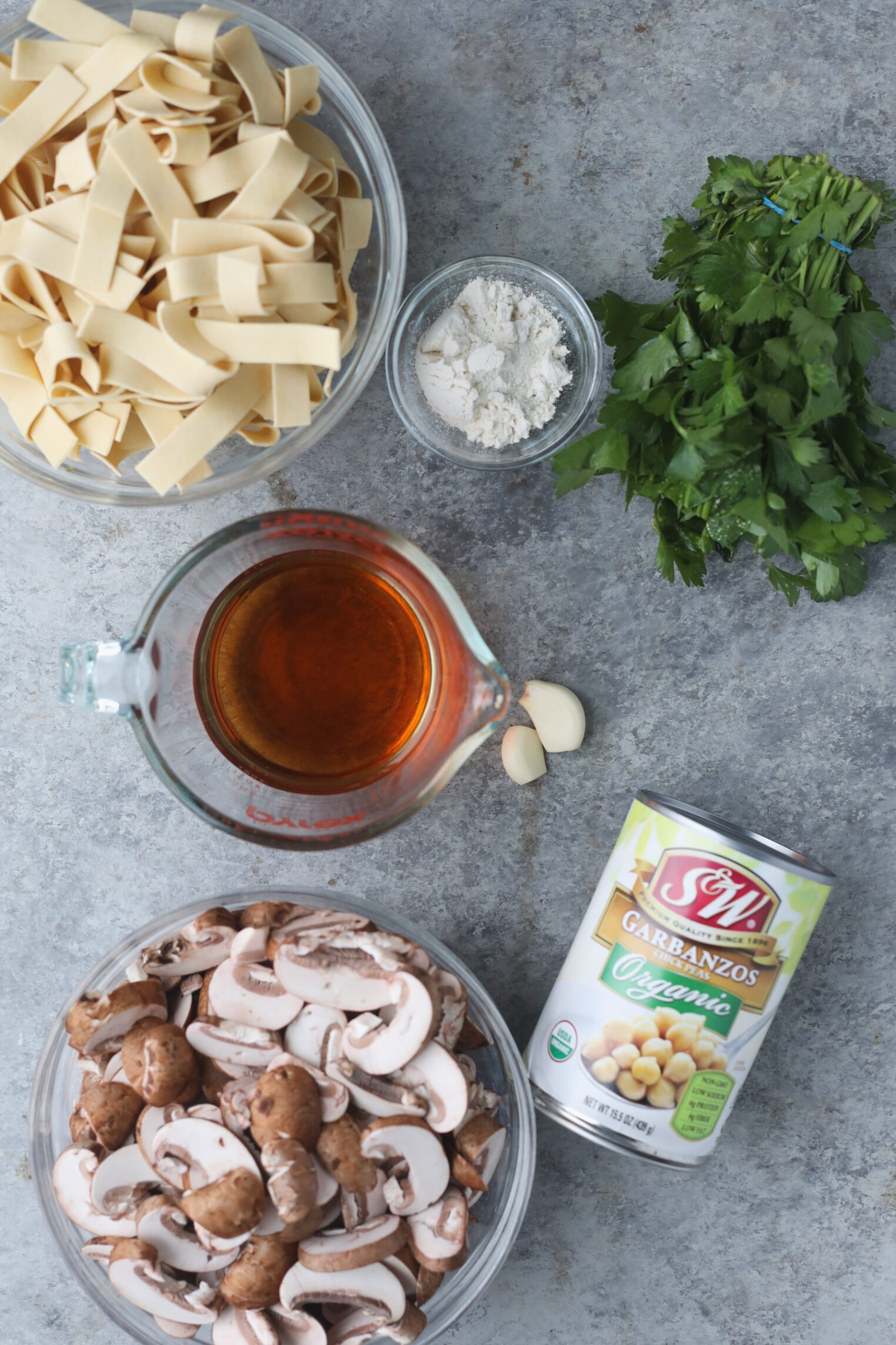 Mushroom Marsala with Chickpeas 2 A Top View Of Recipe Ingredients For Mushroom Marsala On A Gray Surface: Uncooked Pappardelle Pasta, Fresh Parsley, Flour, Vegetable Broth, Garlic Cloves, Sliced Mushrooms, And A Can Of Organic Garbanzo Beans.