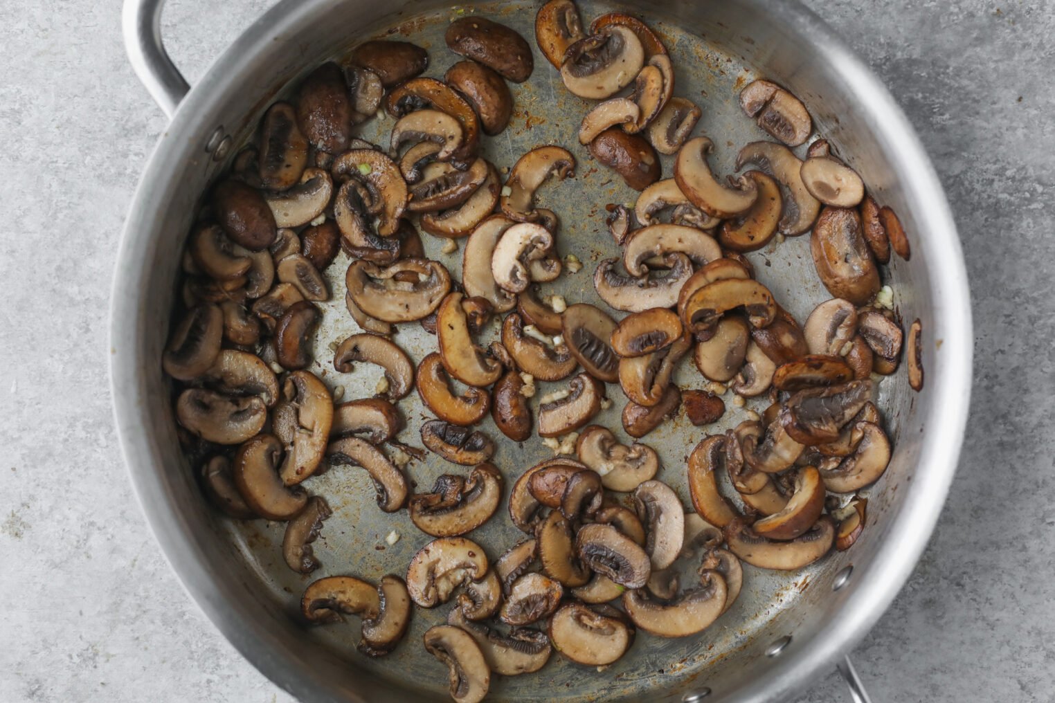 Mushroom Marsala with Chickpeas 5 Sliced Mushrooms For Mushroom Marsala Sautéing In A Large Stainless Steel Pan, Showing A Mix Of Browned And Lightly Cooked Pieces On A Gray Surface.