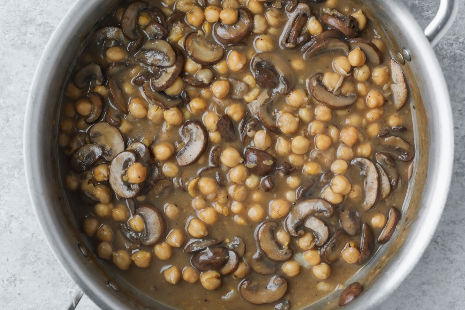 Mushroom Marsala with Chickpeas 6 A Pot Filled With A Hearty Mushroom Marsala Stew, Featuring Sliced Mushrooms And Chickpeas In A Thick Brown Sauce, Viewed From Above On A Light Gray Surface.