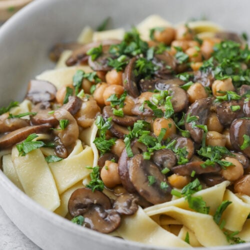 Mushroom Marsala with Chickpeas 10 A Bowl Of Wide Pasta Noodles Topped With Savory Mushroom Marsala, Chickpeas, And Fresh Chopped Parsley, Served On A Light-Colored Surface With Small Bowls Of Herbs In The Background.