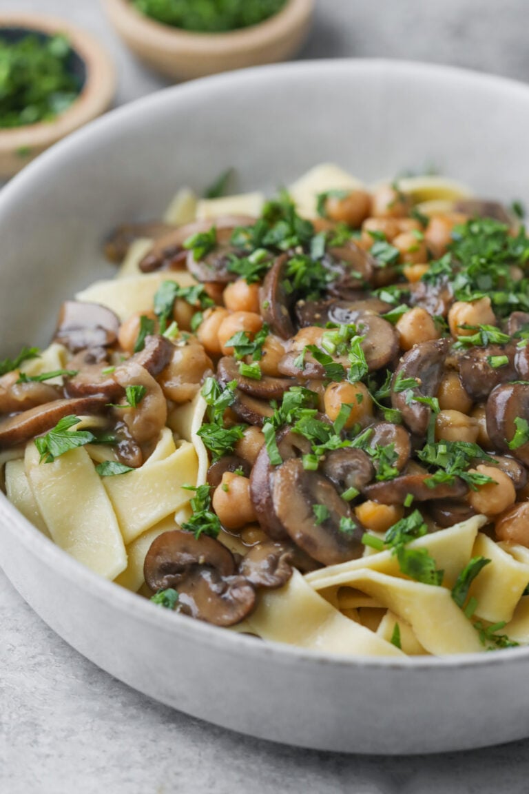 A Bowl Of Wide Pasta Noodles Topped With Savory Mushroom Marsala, Chickpeas, And Fresh Chopped Parsley, Served On A Light-Colored Surface With Small Bowls Of Herbs In The Background.
