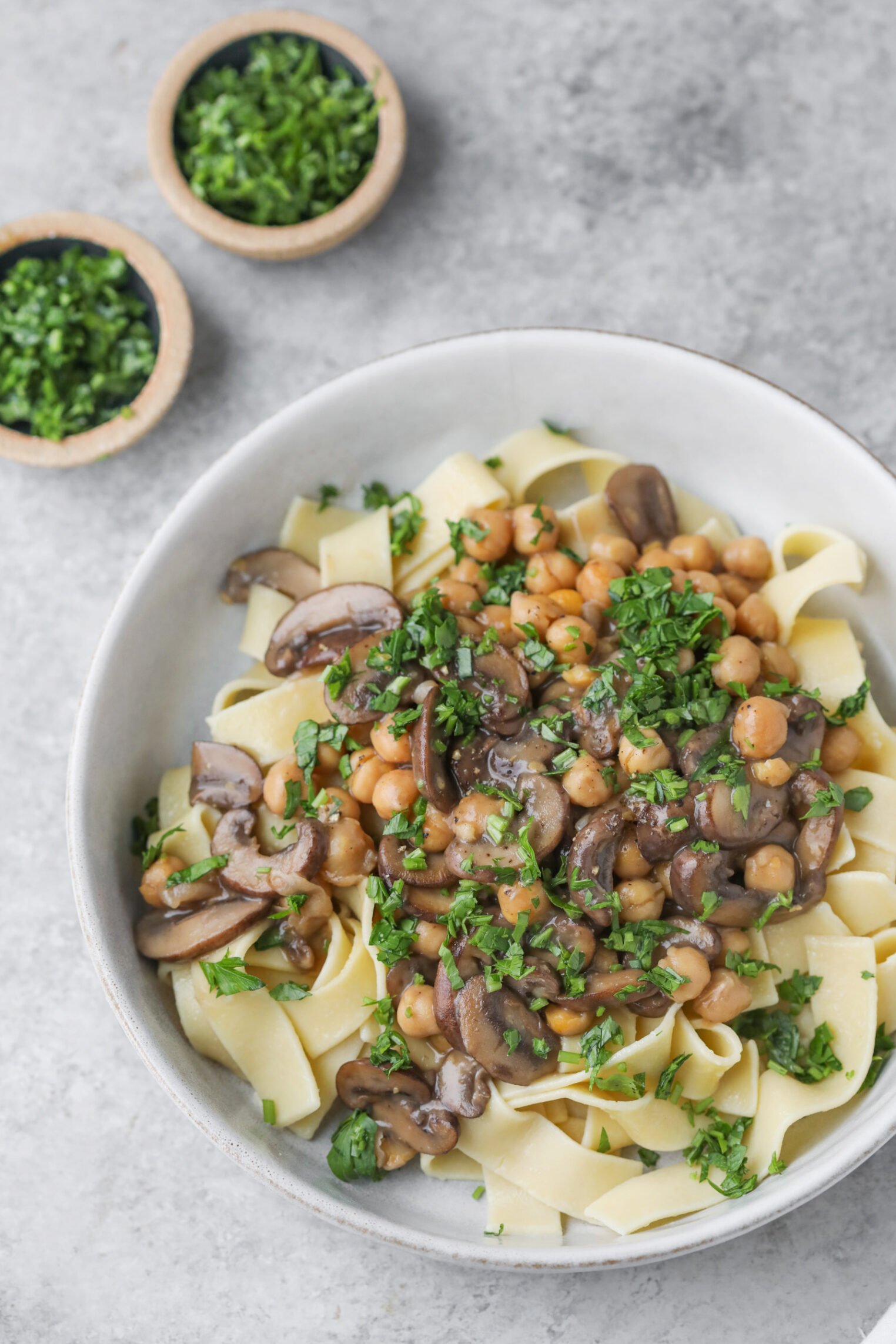 Mushroom Marsala with Chickpeas 3 A Bowl Of Wide Pasta Noodles Topped With A Savory Mushroom Marsala And Chickpea Mixture, Garnished With Chopped Fresh Herbs. Two Small Bowls Of Chopped Herbs Are In The Background On A Gray Surface.