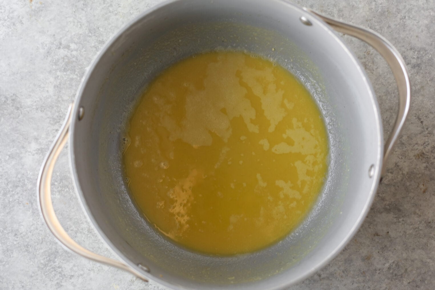 A Top-Down View Of A Gray Metal Pot Containing Melted Butter, With Some Foam On The Surface, Set On A Light Gray Countertop—Perfect For Starting An Okra Stew.