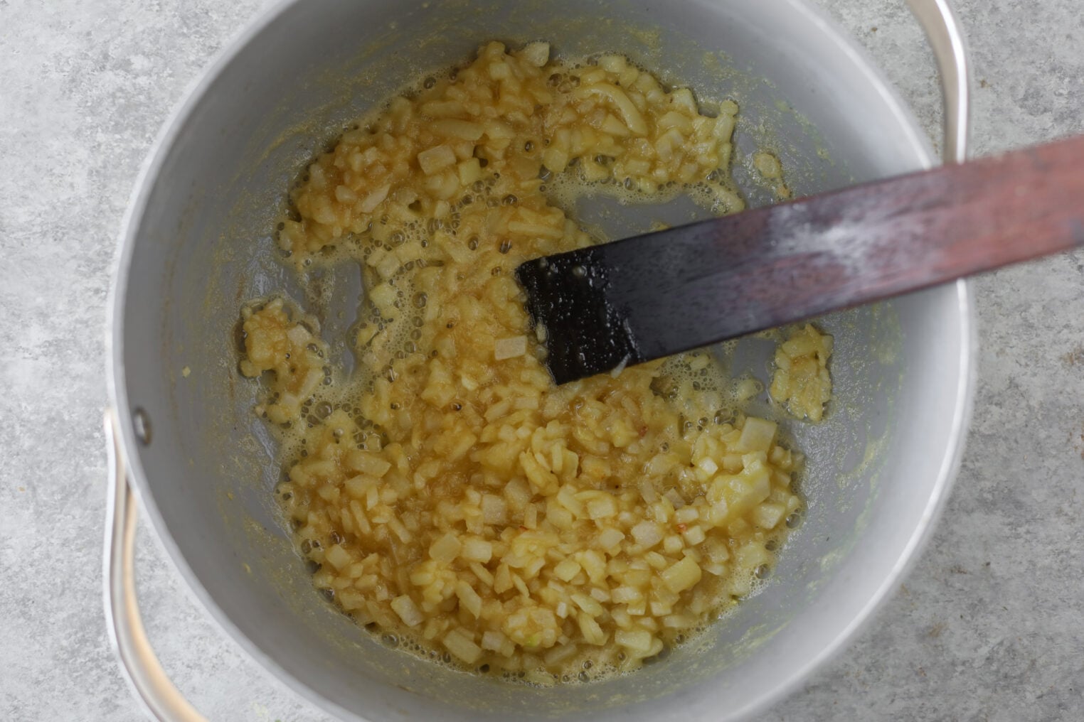 A Pot With Sautéed Diced Onions Being Stirred By A Wooden Spatula, The Perfect Base For Okra Stew. The Soft, Translucent Onions Cook Gently In Oil Or Butter On A Light Gray Surface.