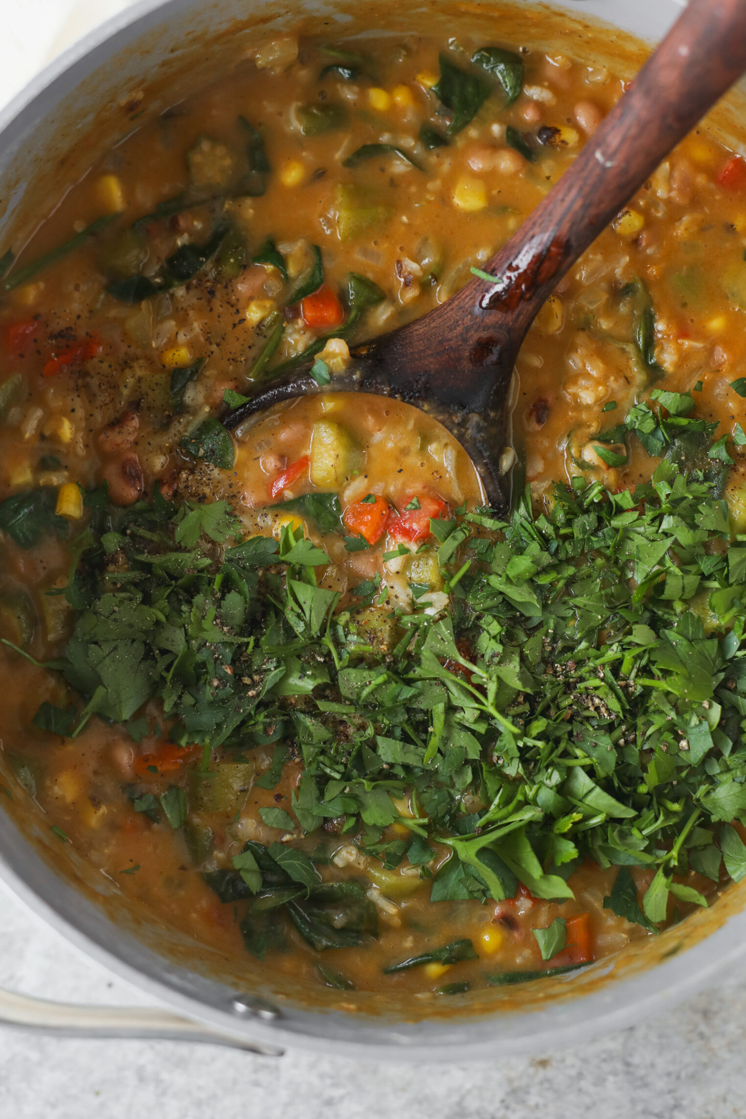 A Pot Of Hearty Vegetable Soup, Reminiscent Of Okra Stew, With Chopped Greens, Corn, Beans, And Diced Red And Green Vegetables Is Being Stirred With A Wooden Spoon. Fresh Herbs Are Sprinkled On Top.