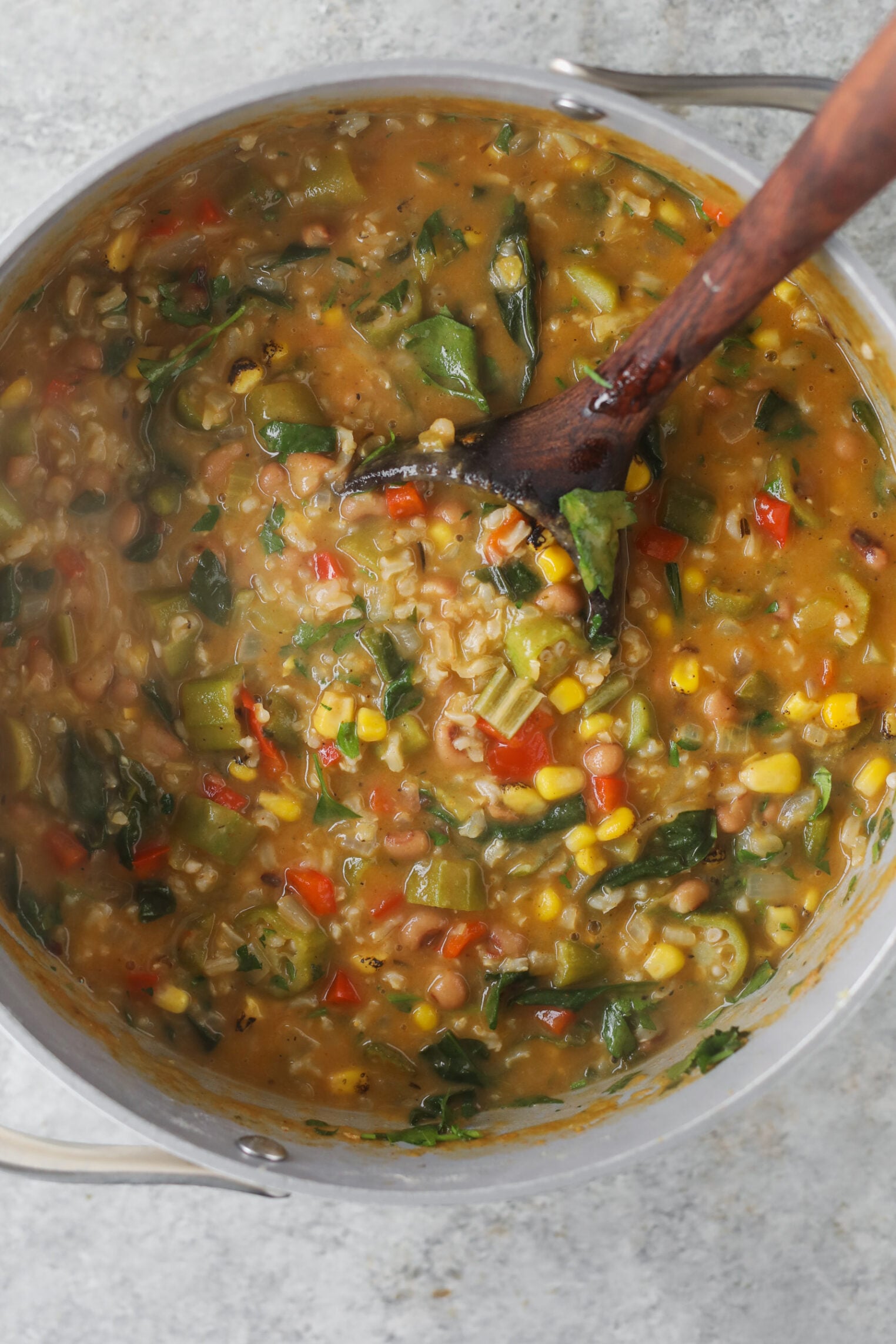 A Large Pot Filled With A Hearty Okra Stew Featuring Corn, Beans, Spinach, And Diced Peppers Is Being Stirred With A Wooden Spoon.