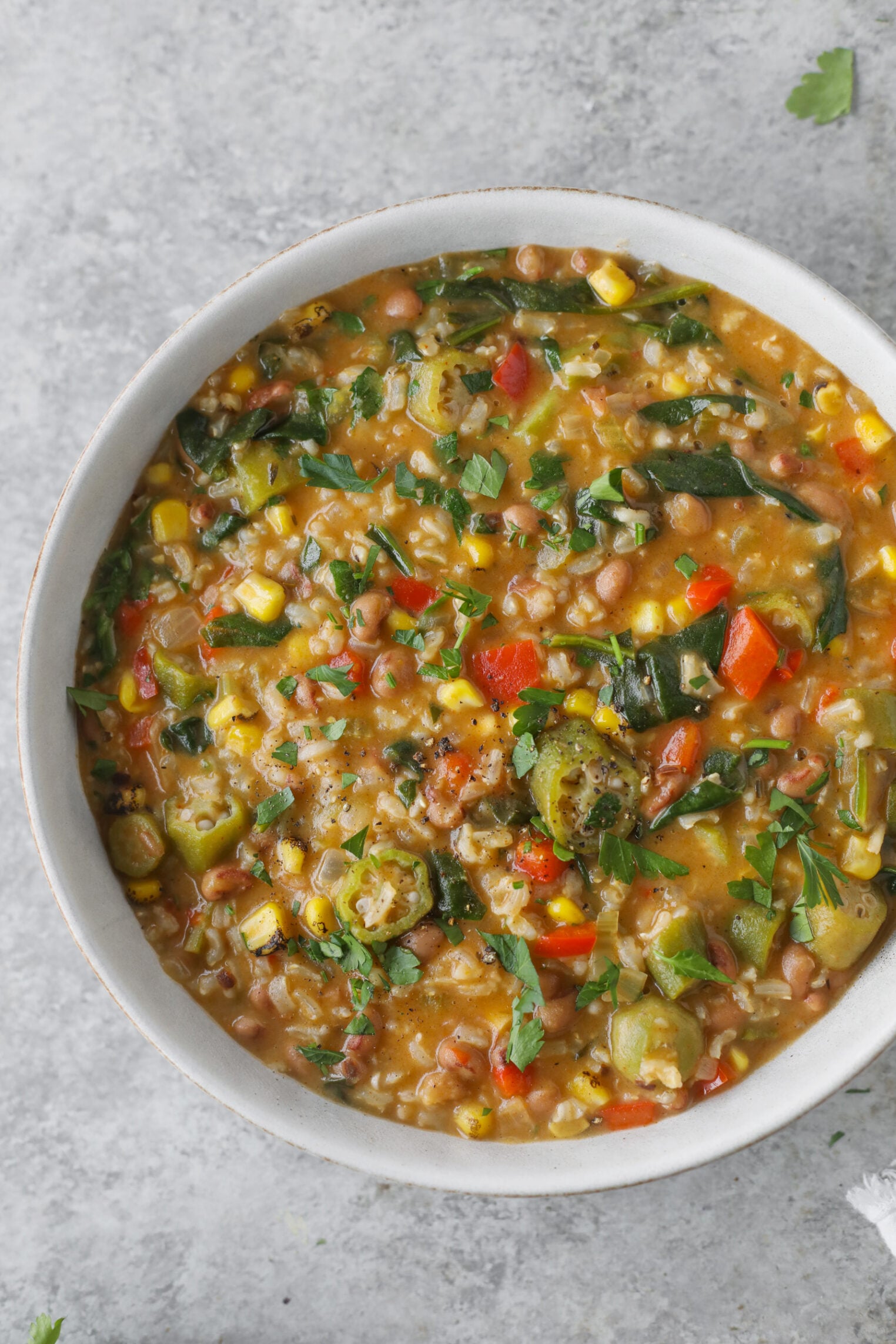 A Bowl Of Hearty Okra Stew Brimming With Corn, Beans, Chopped Greens, Red Bell Peppers, And Herbs, Set On A Light Gray Surface.