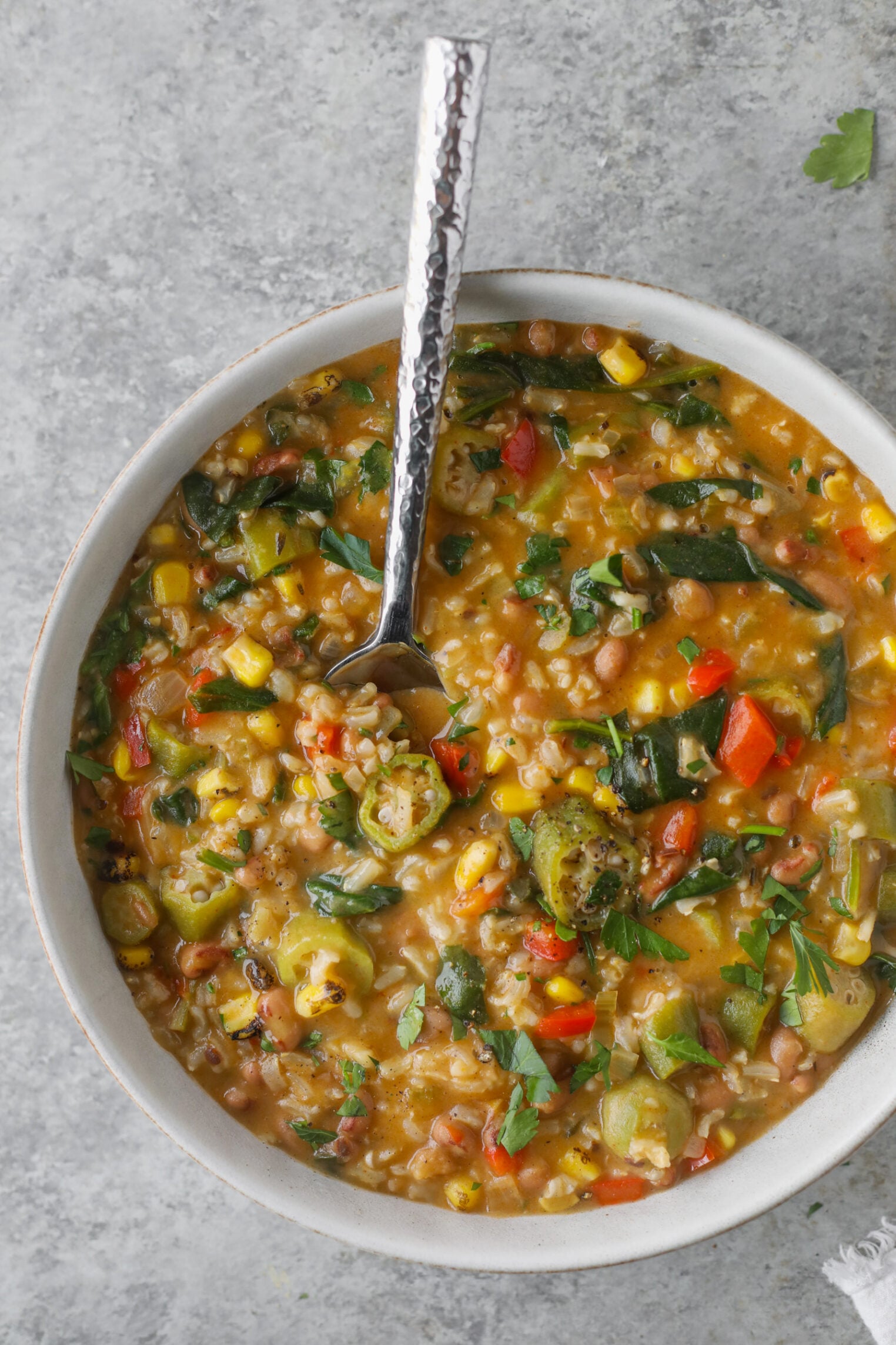 A Bowl Of Hearty Okra Stew With Corn, Spinach, Red Bell Peppers, And Grains, Garnished With Fresh Herbs, Sits On A Gray Surface With A Metal Spoon In The Bowl.