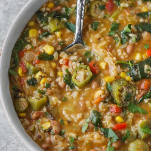 A Bowl Of Vegetable Gumbo, Reminiscent Of A Classic Okra Stew, With Corn, Beans, Rice, Peppers, And Fresh Herbs. A Spoon Rests Inside The Bowl On A Light Grey Surface.
