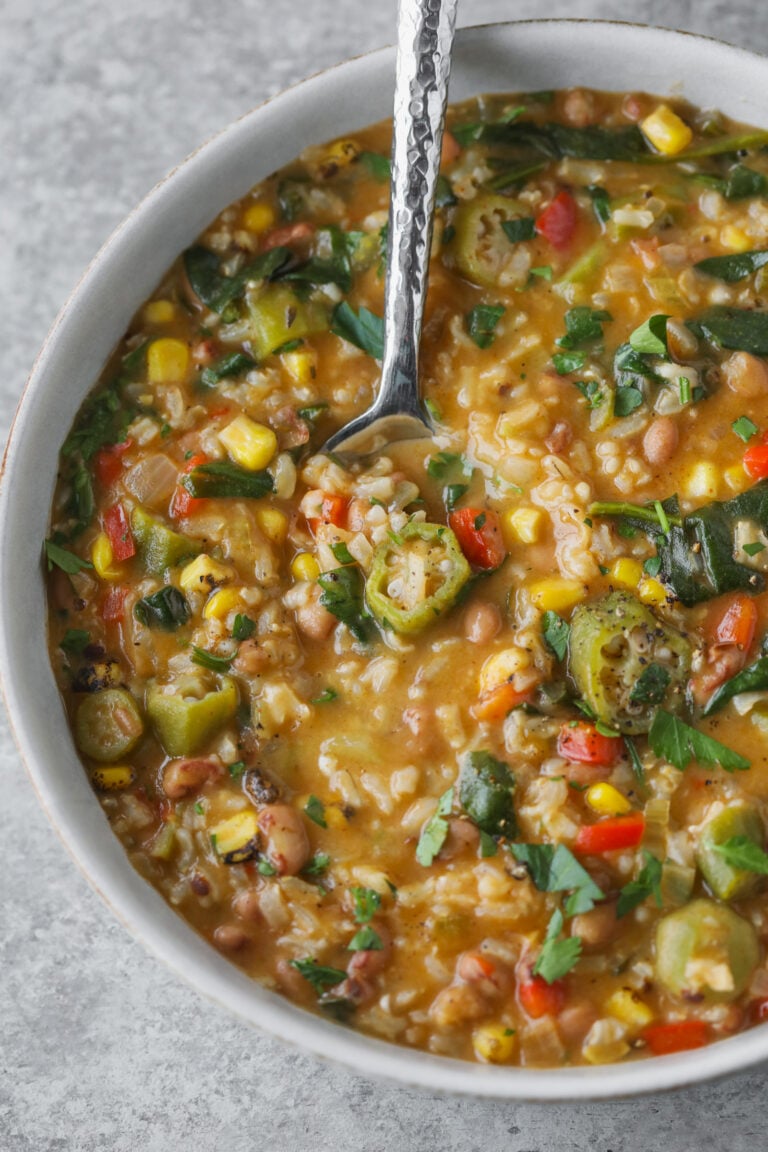 A Bowl Of Vegetable Gumbo, Reminiscent Of A Classic Okra Stew, With Corn, Beans, Rice, Peppers, And Fresh Herbs. A Spoon Rests Inside The Bowl On A Light Grey Surface.
