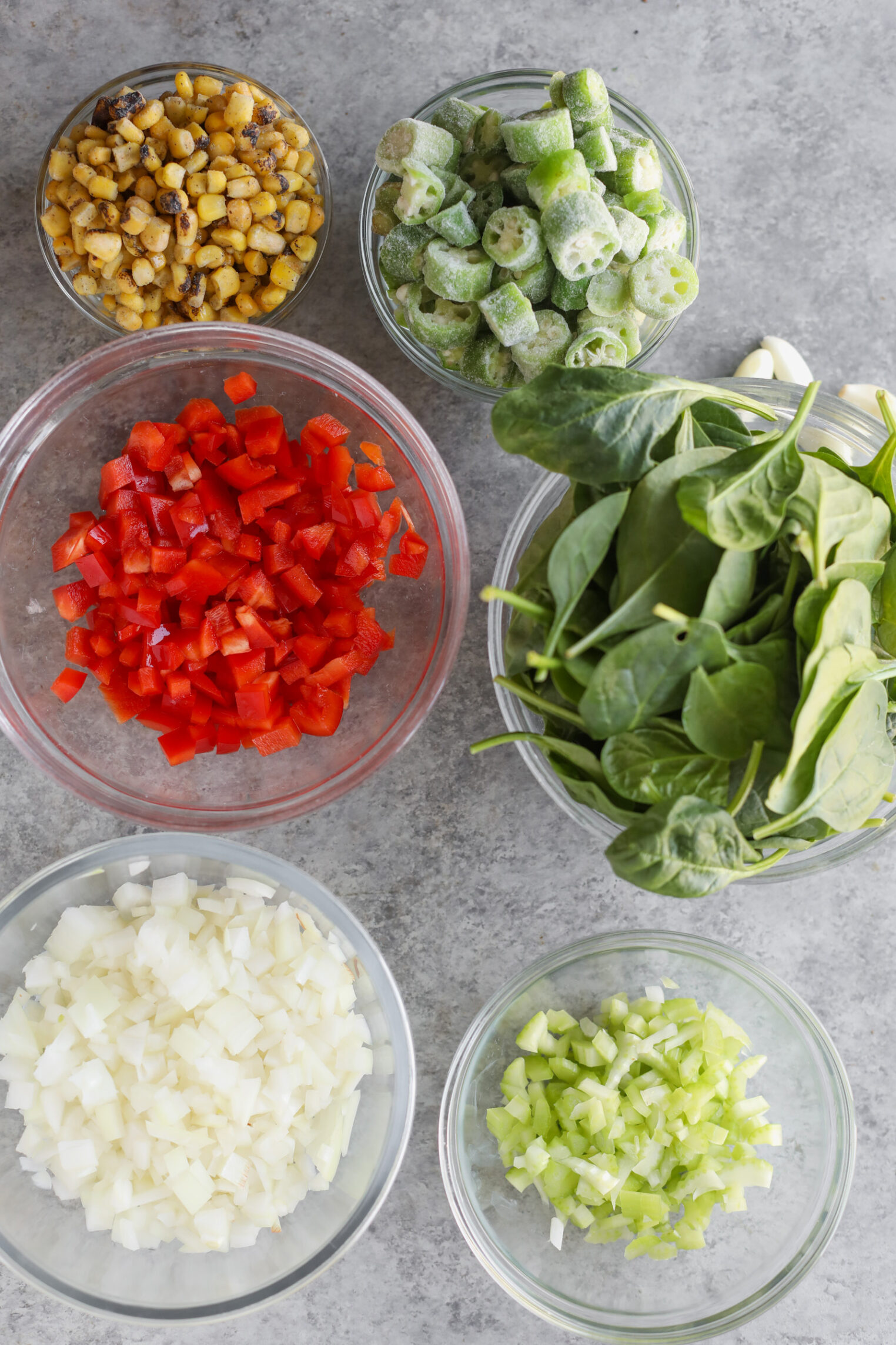 Several Glass Bowls Filled With Chopped Vegetables—Including Red Bell Pepper, Onion, Celery, Spinach Leaves, Frozen Okra For Okra Stew, And Roasted Corn—Are Arranged On A Gray Countertop.