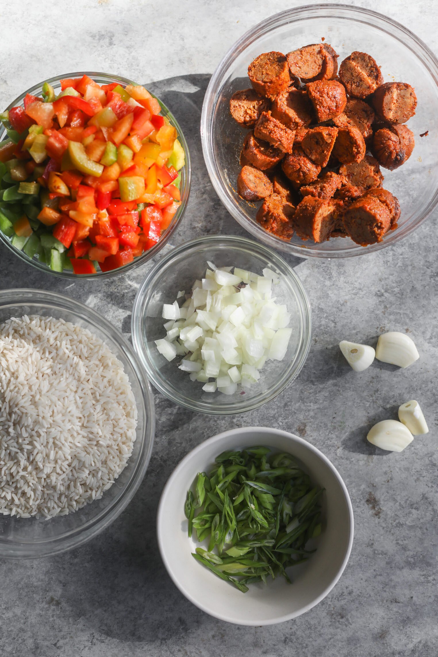 Five Bowls With Chopped Ingredients On A Gray Surface: Diced Colorful Bell Peppers, Onions, Uncooked White Rice, Sliced Green Onions, And Vegan Sausage—Perfect For Assembling A Vibrant Vegan Jambalaya. Three Whole Garlic Cloves Sit Nearby.