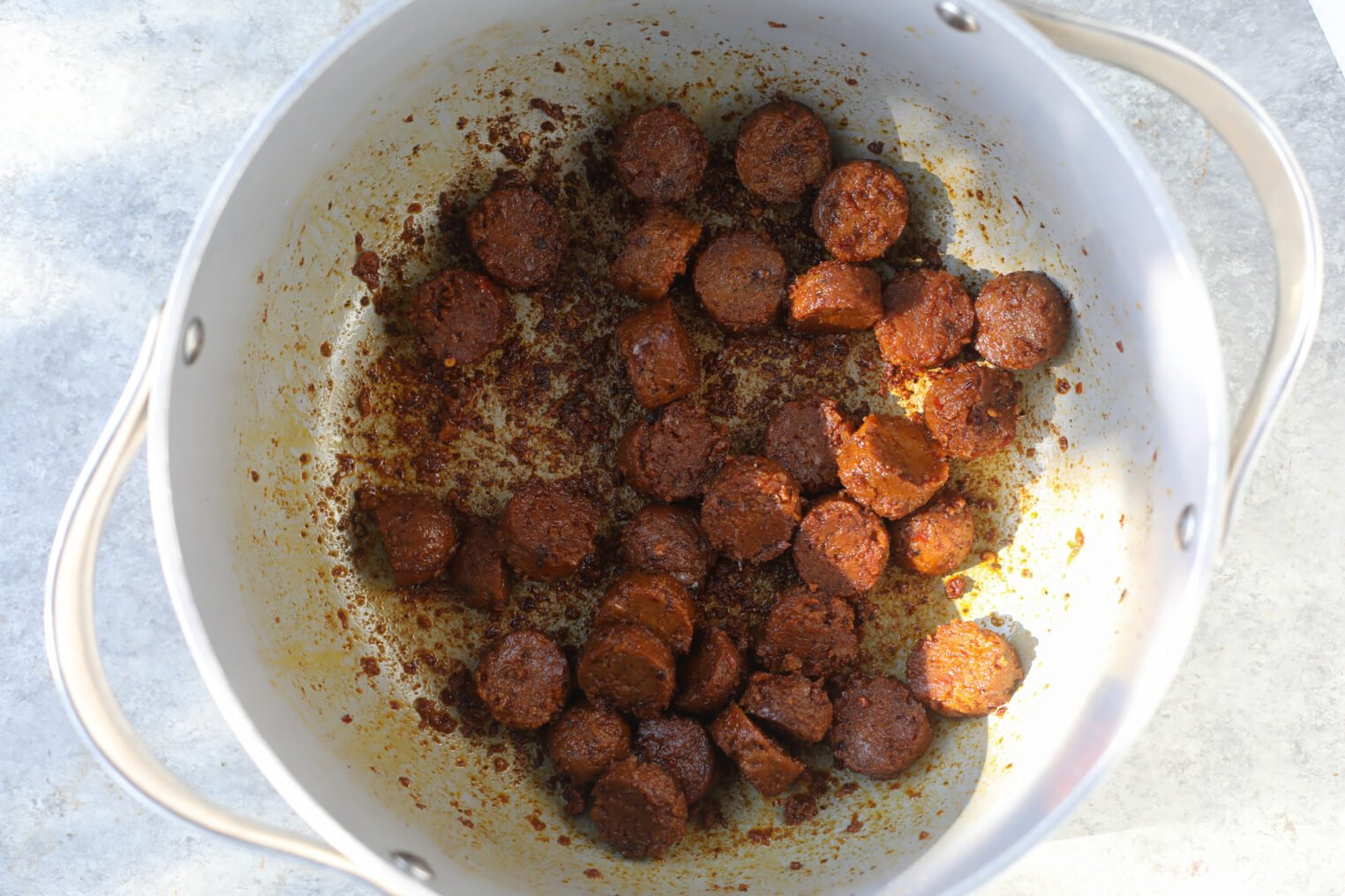 Sliced Sausage Pieces Browned With Spices In A White Pot, Seen From Above On A Light Surface—Perfect For Adding To Vegan Jambalaya.