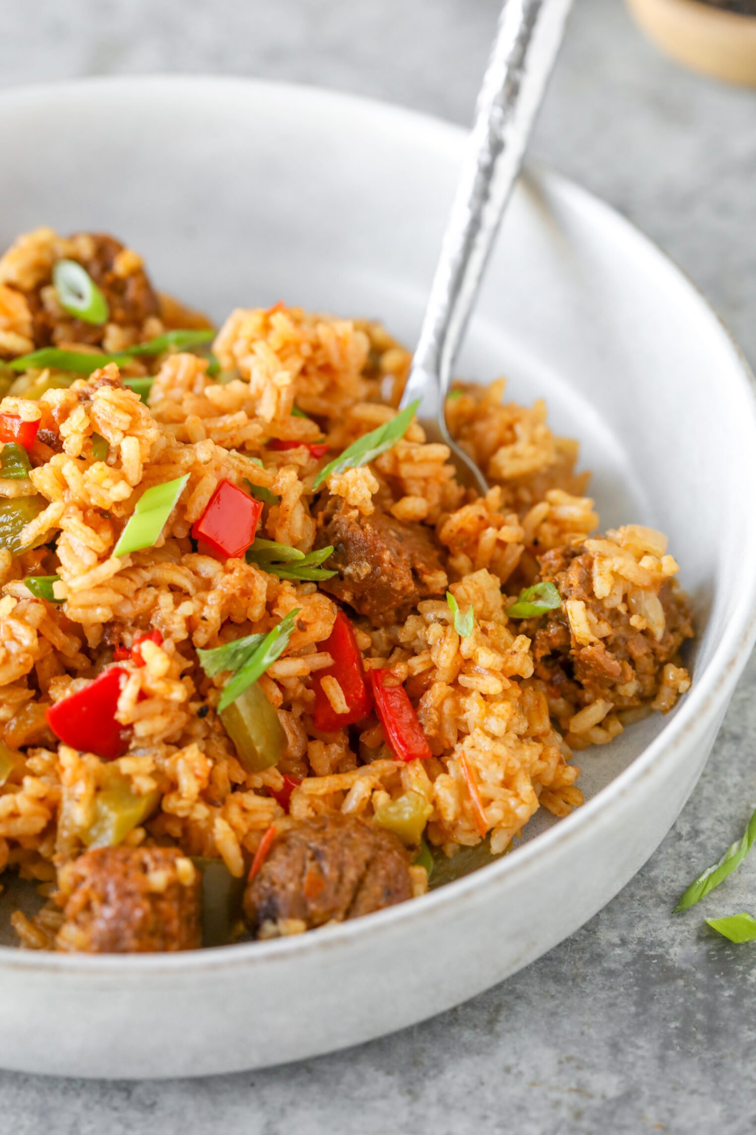 A Bowl Of Seasoned Rice Mixed With Chunks Of Sausage, Red And Green Bell Peppers, And Garnished With Sliced Green Onions, This Vegan Jambalaya Invites You To Dig In With A Fork Resting In The Bowl.