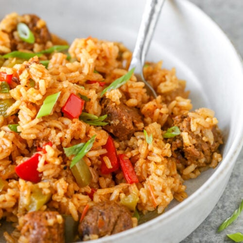 A Bowl Of Seasoned Rice Mixed With Chunks Of Sausage, Red And Green Bell Peppers, And Garnished With Sliced Green Onions, This Vegan Jambalaya Invites You To Dig In With A Fork Resting In The Bowl.