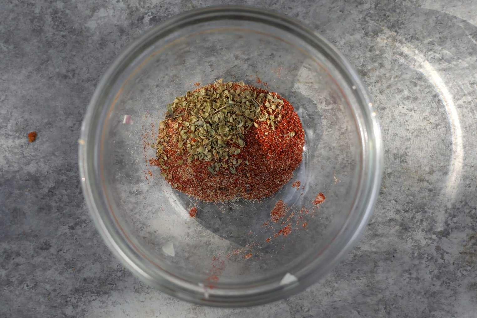 A Glass Jar Containing A Colorful Mixture Of Dried Spices, Perfect For Vegan Jambalaya, Including Greenish Dried Herbs And Reddish Powder, Sits On A Gray Textured Surface.