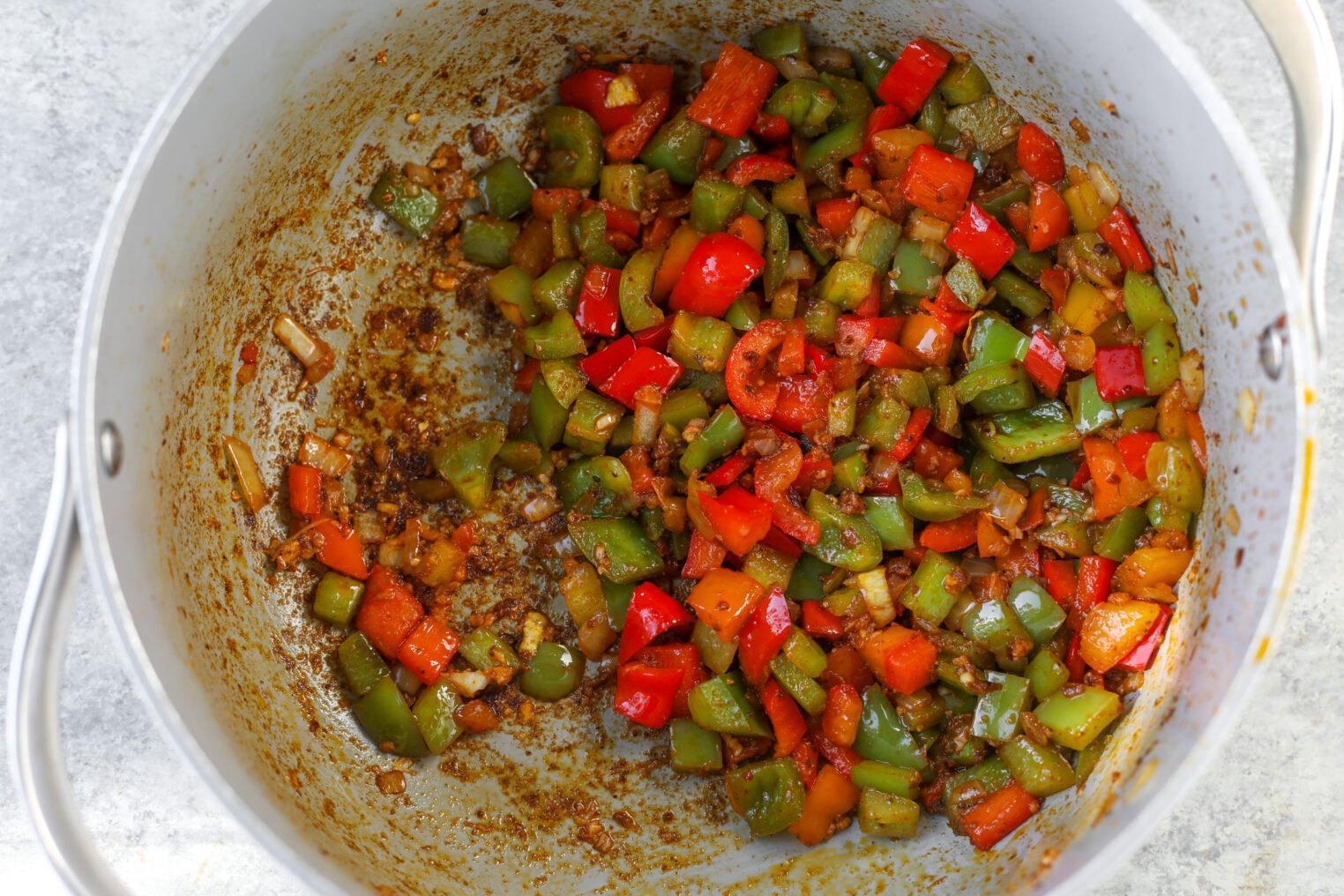 Diced Green And Red Bell Peppers With Onions Are Sautéed In A Large Silver Pot, Showing Browned Seasoning And Caramelization—The Perfect Base For A Flavorful Vegan Jambalaya.