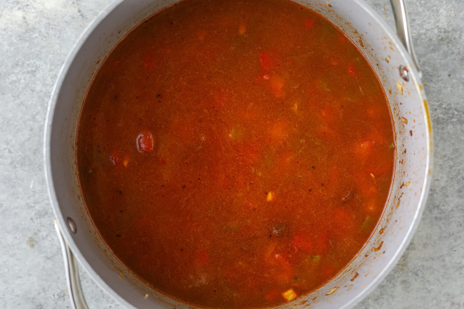 A Pot Of Red-Brown Broth-Based Vegan Jambalaya Soup With Visible Diced Vegetables, Such As Peppers And Tomatoes, Sits On A Gray Countertop.