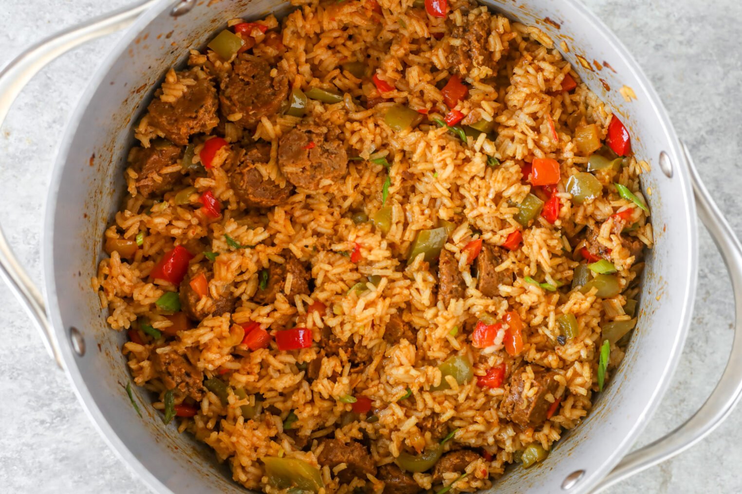 A Large Pot Filled With Vegan Jambalaya, Featuring Cooked Rice, Sliced Sausage, Chopped Green And Red Bell Peppers, And Savory Seasonings. The Colorful Dish Is Packed With Visible Spices And Vibrant Vegetables.