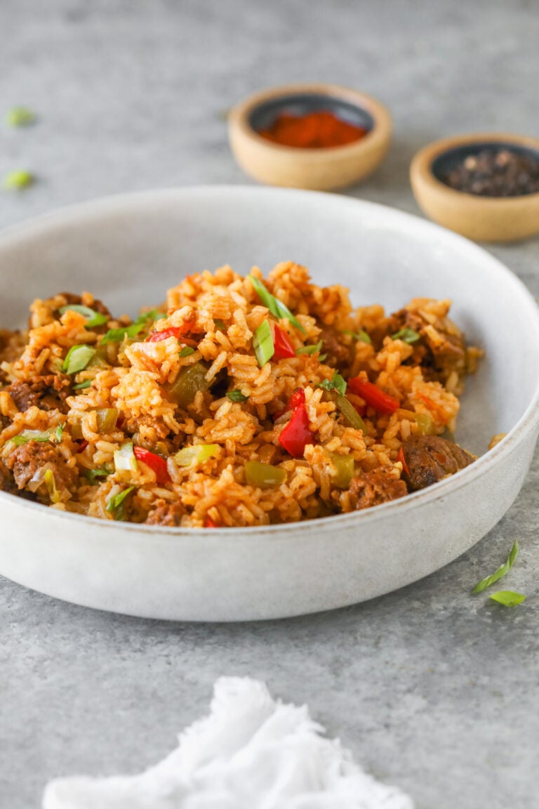 A White Bowl Filled With Vegan Jambalaya—Seasoned Rice, Plant-Based Meat, And Chopped Red And Green Bell Peppers, Garnished With Green Onions. Two Small Spice Bowls Are Blurred In The Background On A Gray Surface.