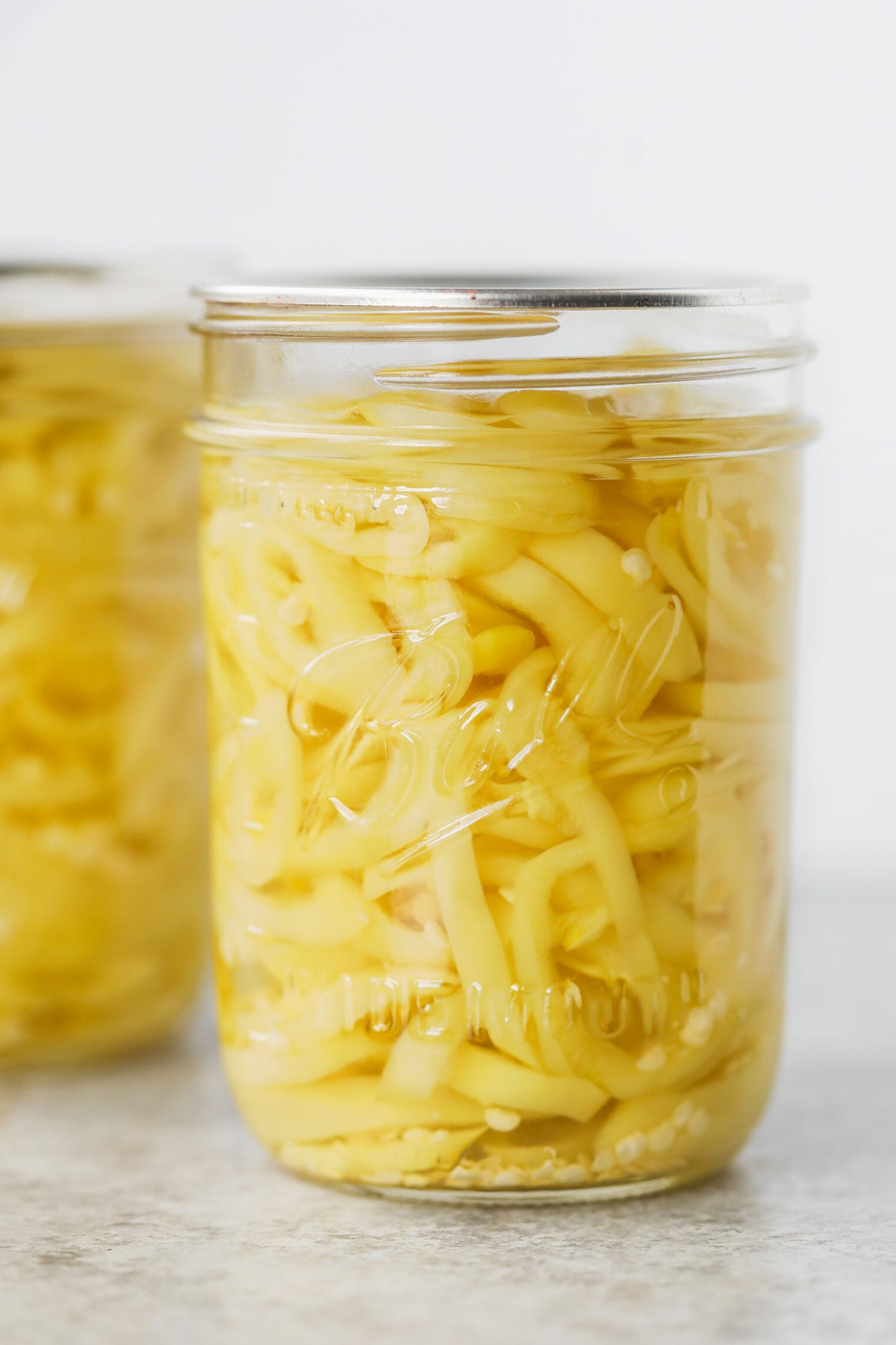 Pickled Jalapeños (Canning Recipe) 9 A Glass Jar Filled With Sliced Yellow Banana Peppers In Liquid, Sealed With A Metal Lid, Sits On A Light-Colored Surface. Another Similar Jar—This One Featuring Pickled Jalapenos—Is Partially Visible In The Background.