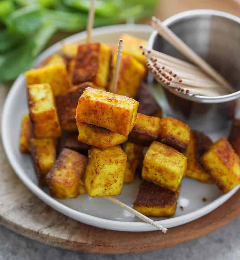 Close-Up Of A White Plate Showcasing Skewered Paneer Cubes, Golden Brown And Crispy, Seasoned With Spices—Delicious, Healthy Appetizers. A Small Metal Cup With Wooden Toothpicks Sits Beside Them As Fresh Greens Blur In The Background.