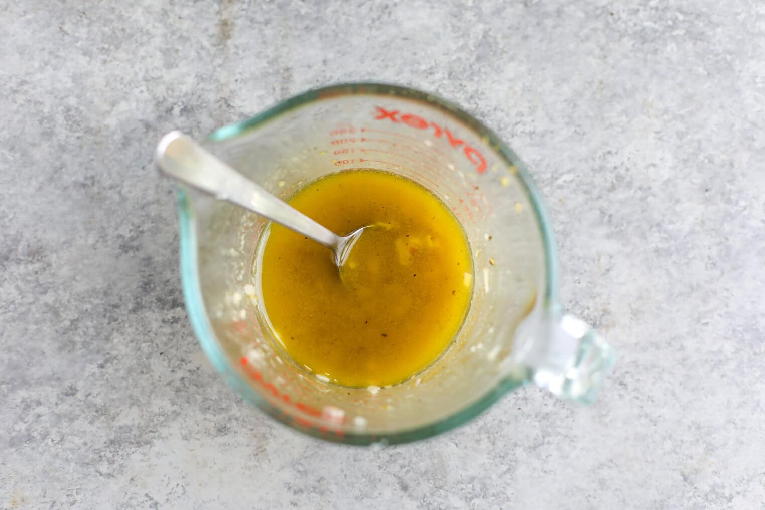 Shaved Asparagus Spring Salad 6 A Glass Measuring Cup With Yellow Liquid And A Metal Spoon Inside, Sitting On A Gray Countertop. The Liquid Appears To Be A Dressing Or Sauce, Perfect For Drizzling Over A Shaved Asparagus Spring Salad.