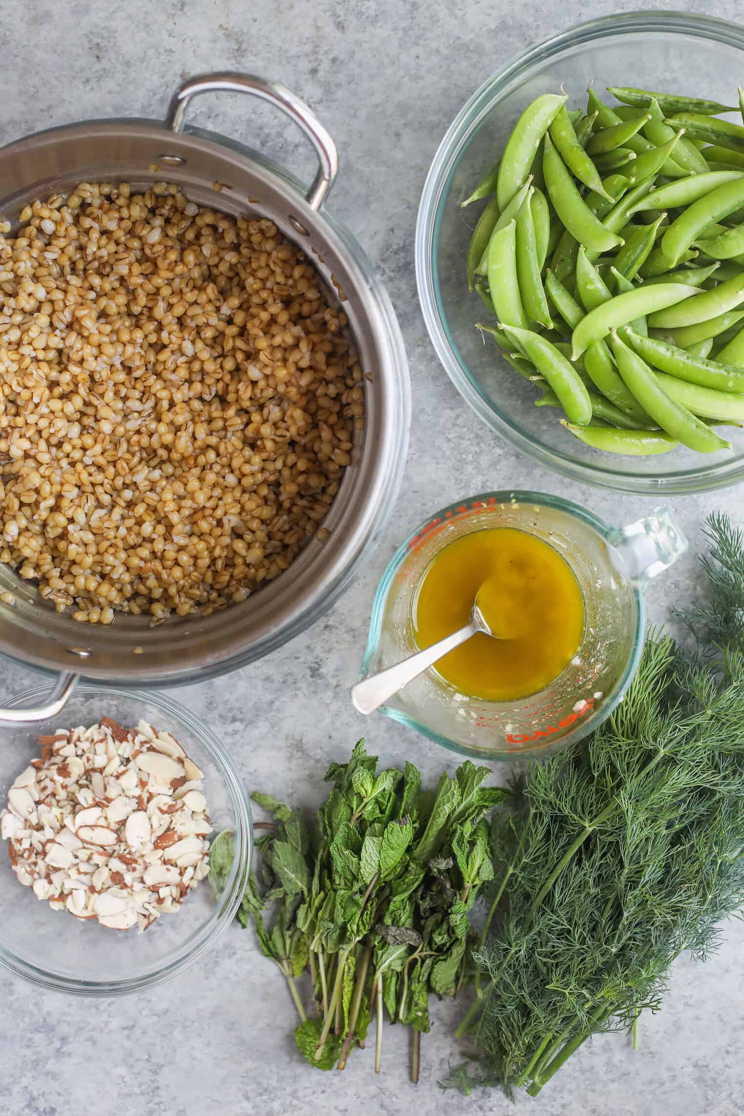 Spring Pea, Mint and Wheat Berry Salad 2 Ingredients For A Spring Pea Salad On A Gray Surface: A Pot Of Cooked Wheat Berries, A Bowl Of Snap Peas, Chopped Almonds, Fresh Mint, Fresh Dill, And A Glass Measuring Cup With Yellow Dressing And A Spoon.