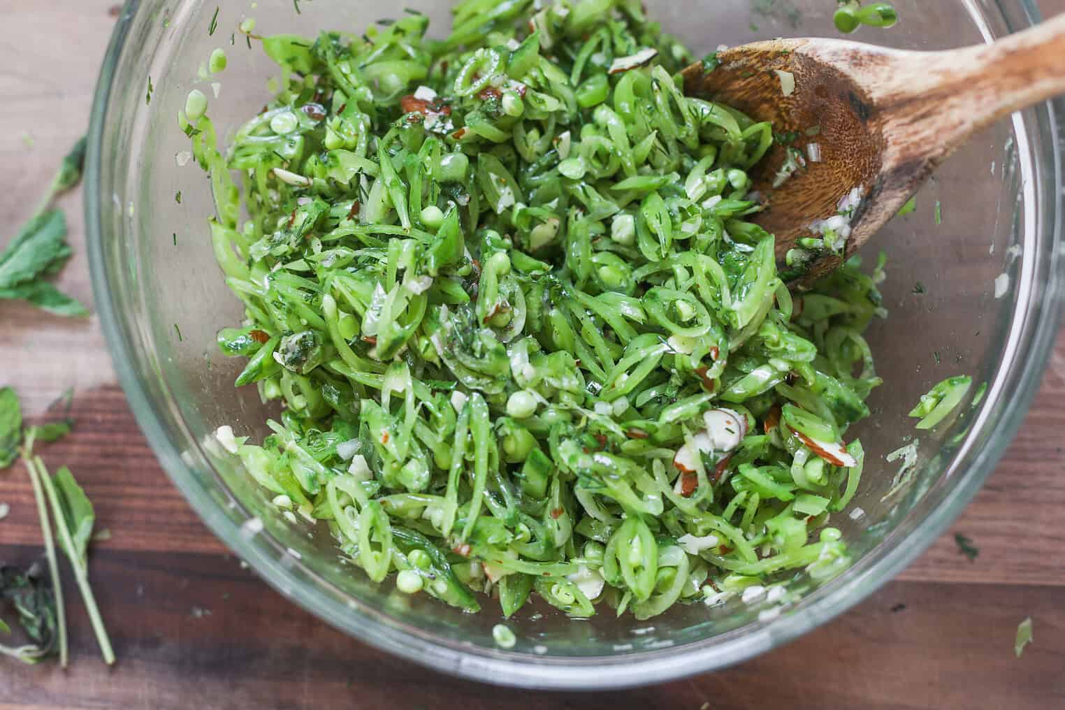 Spring Pea, Mint and Wheat Berry Salad 7 A Glass Bowl Filled With A Fresh Spring Pea Salad Made Of Finely Sliced Snap Peas, Herbs, And Seasonings. A Wooden Spoon Rests In The Bowl, And The Mixture Looks Crisp And Vibrant On A Wooden Surface.