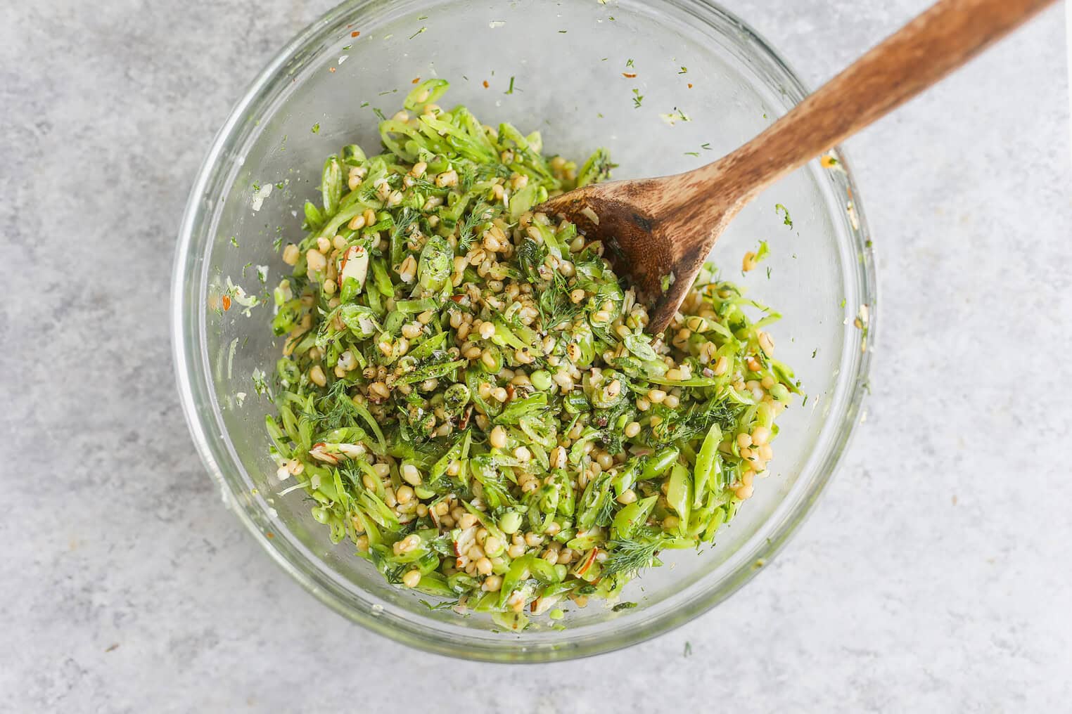 Spring Pea, Mint and Wheat Berry Salad 8 A Glass Bowl Filled With A Fresh Spring Pea Salad Featuring Chopped Herbs, Grains, And Seeds, Mixed With A Wooden Spoon And Sitting On A Light Gray Surface.