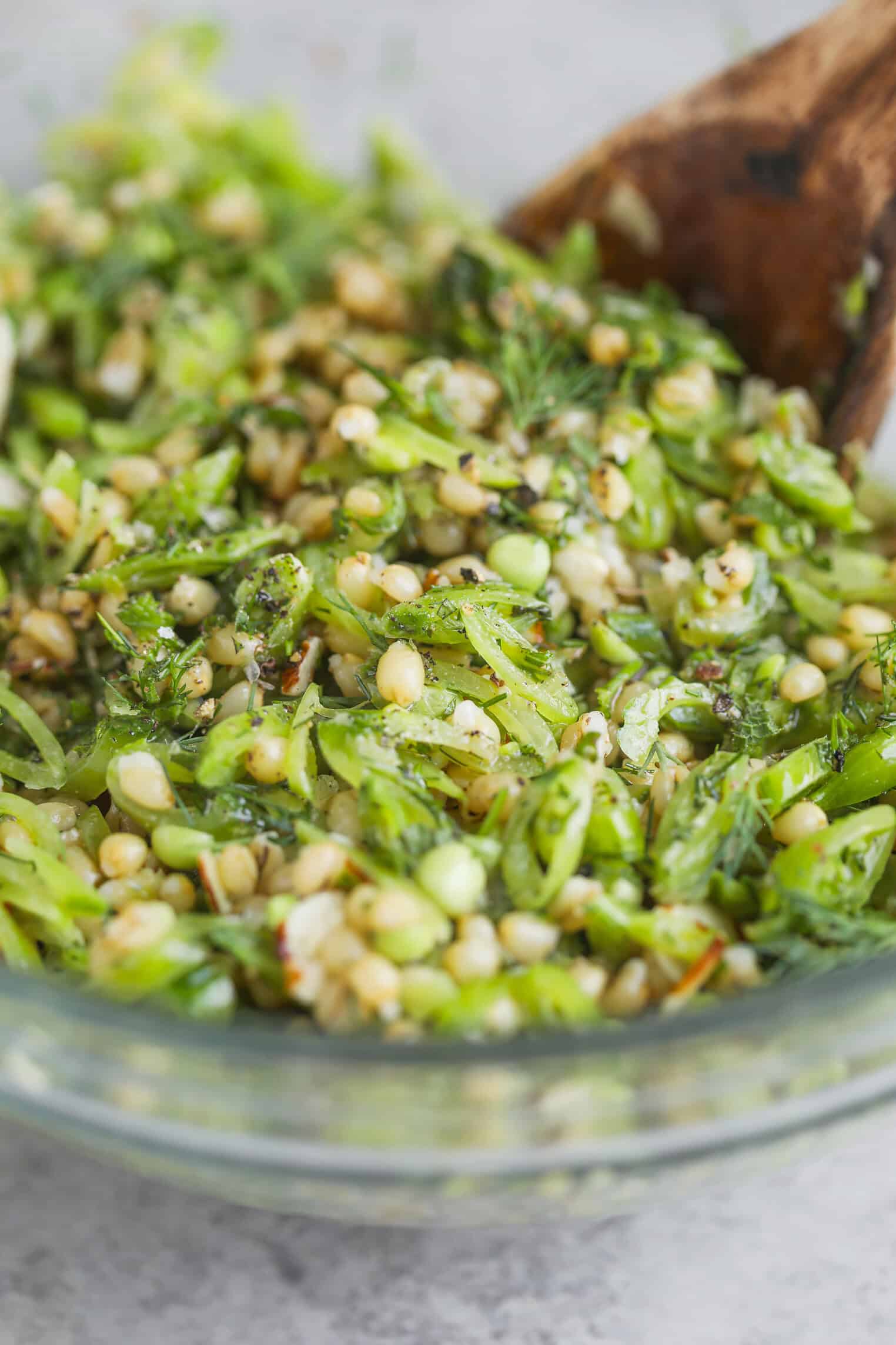 Spring Pea, Mint and Wheat Berry Salad 9 A Close-Up Of A Glass Bowl Filled With A Fresh Spring Pea Salad Made Of Chopped Greens, Herbs, Nuts, And Seeds, With A Wooden Spoon Partially Visible In The Mixture.