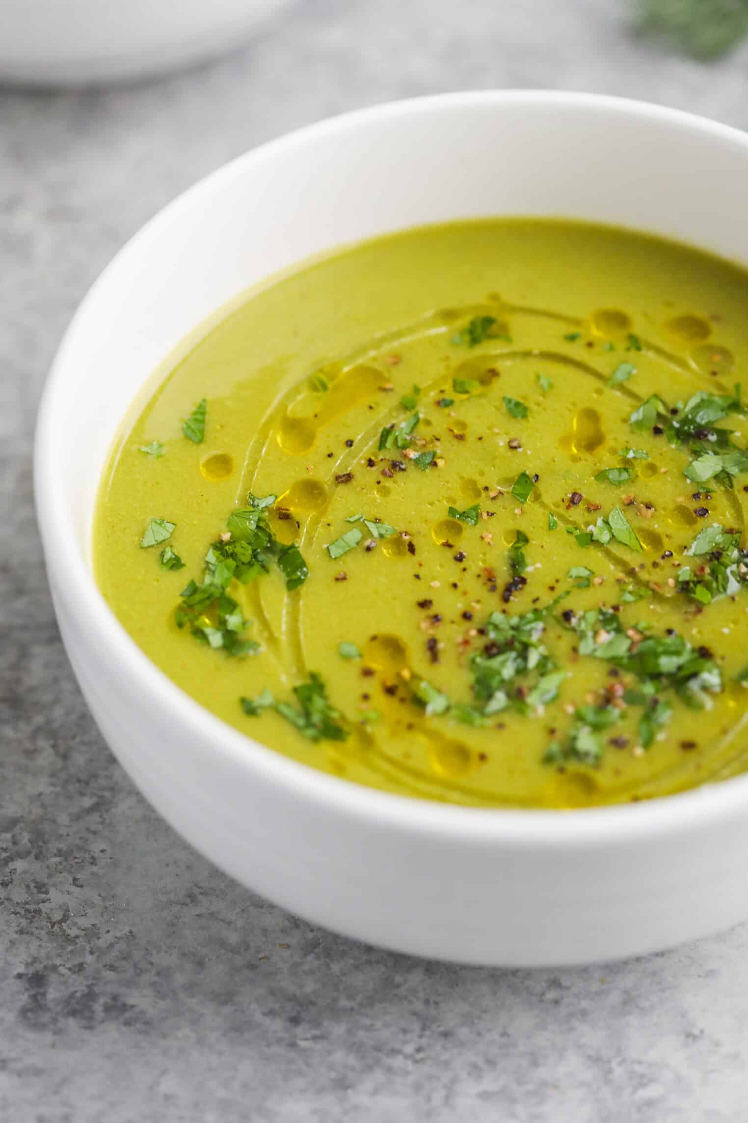 A White Bowl Filled With Creamy Pea And Asparagus Soup, Garnished With Chopped Herbs, Cracked Black Pepper, And A Drizzle Of Olive Oil, Sits On A Light Gray Surface.